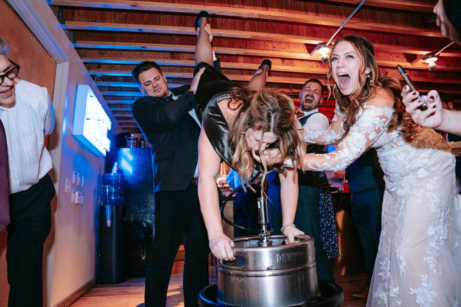 Bride doing keg stand surrounded by cheering wedding guests at lively reception.