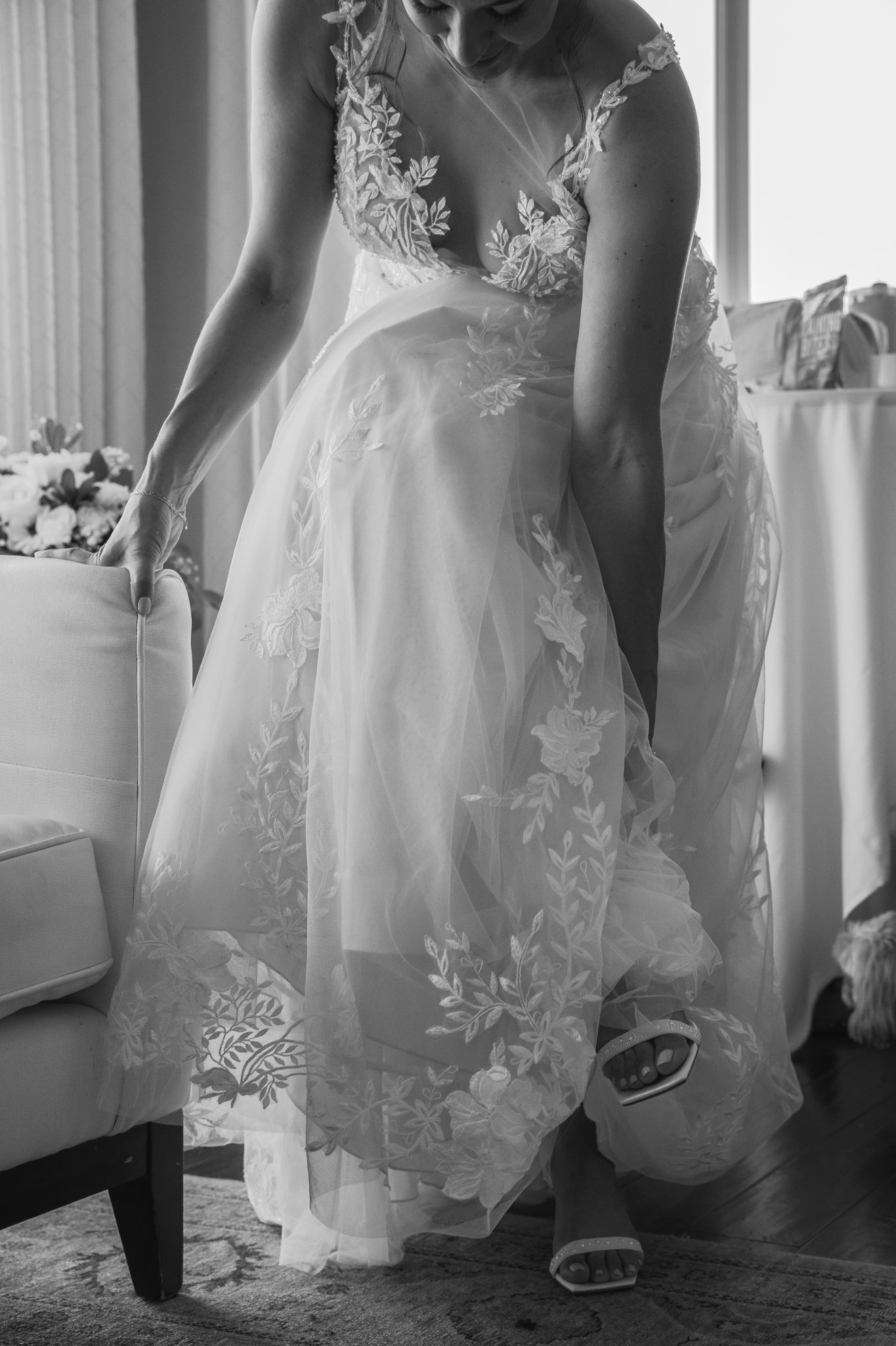 Black and white photo of a bride in a floral lace wedding gown adjusting her dress indoors.