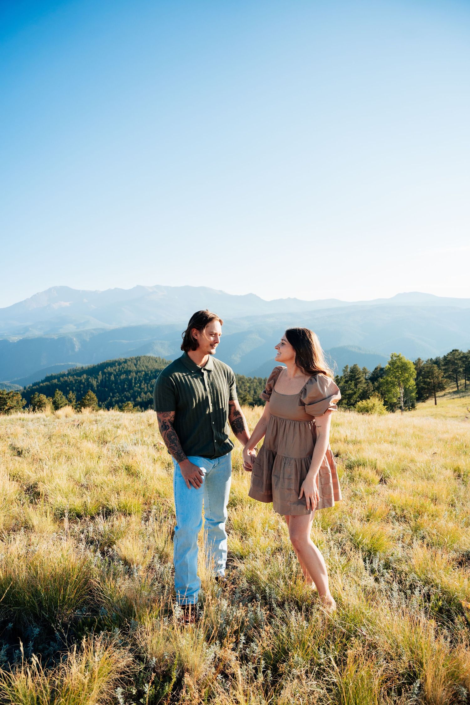 Couple holding hands and smiling at each other in a golden grass field with majestic mountain views behind them.