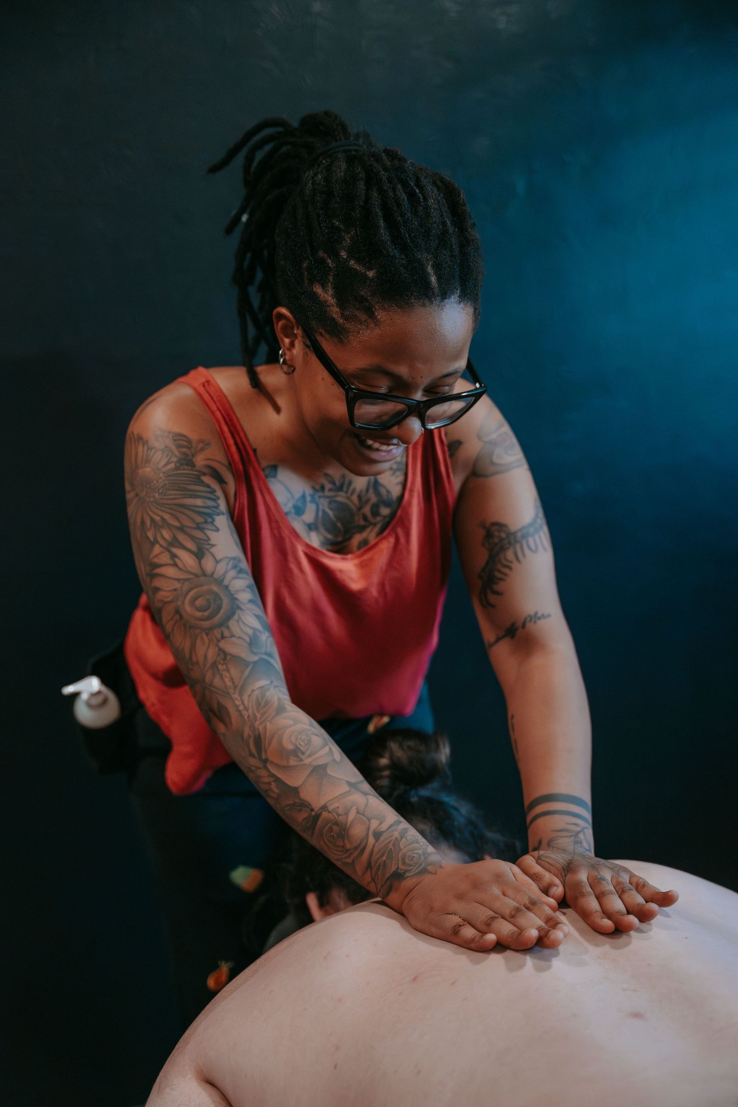 Heavily tattooed female massage therapist smiling while applying pressure to a client's back during a session.