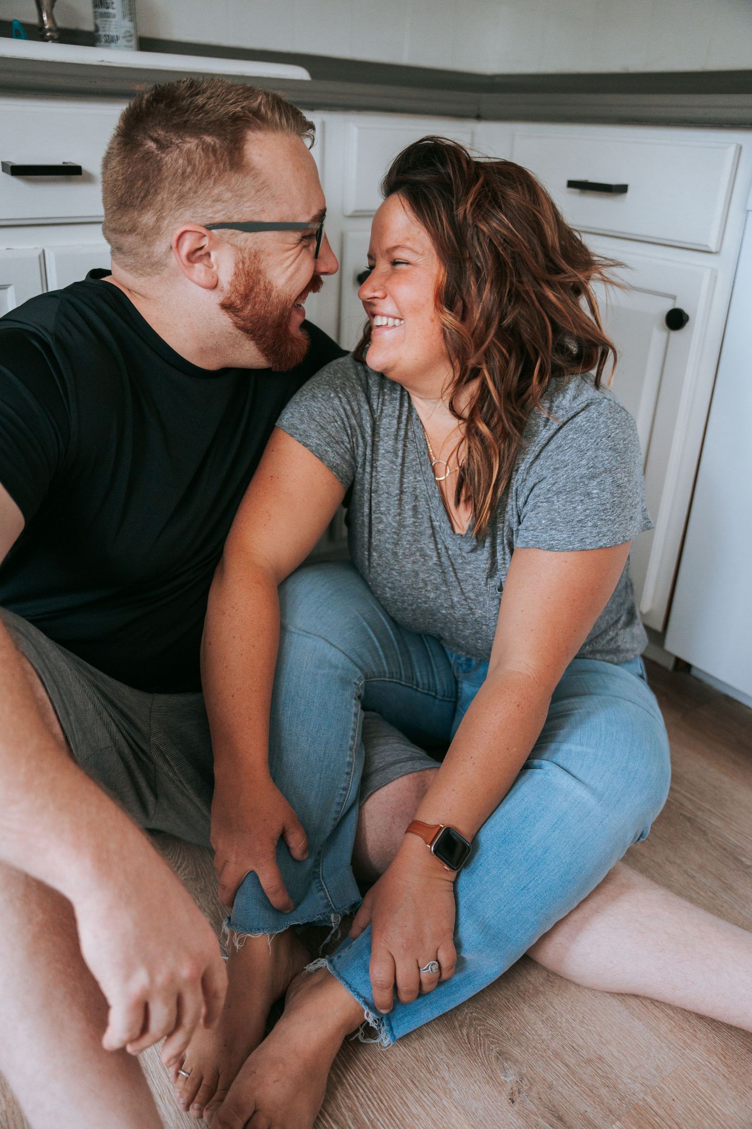 A laughing couple sits together on a kitchen floor gazing at each other during a cozy indoor engagement shoot.