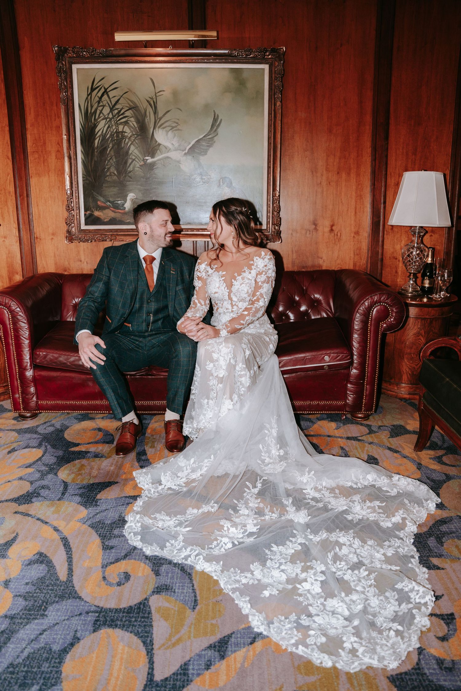 Bride in floral lace gown with long train sits with groom in green suit on red leather sofa.