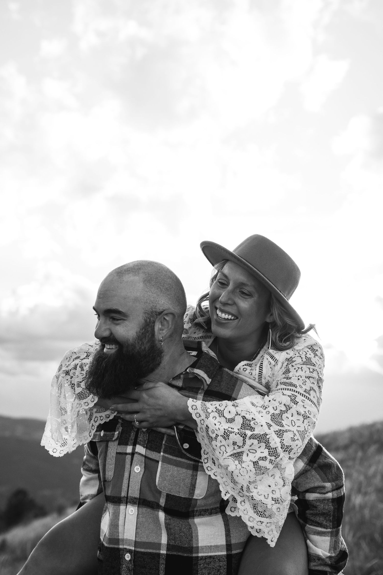 Black and white photo of smiling bride in lace dress and hat getting piggyback ride from bearded groom outdoors.