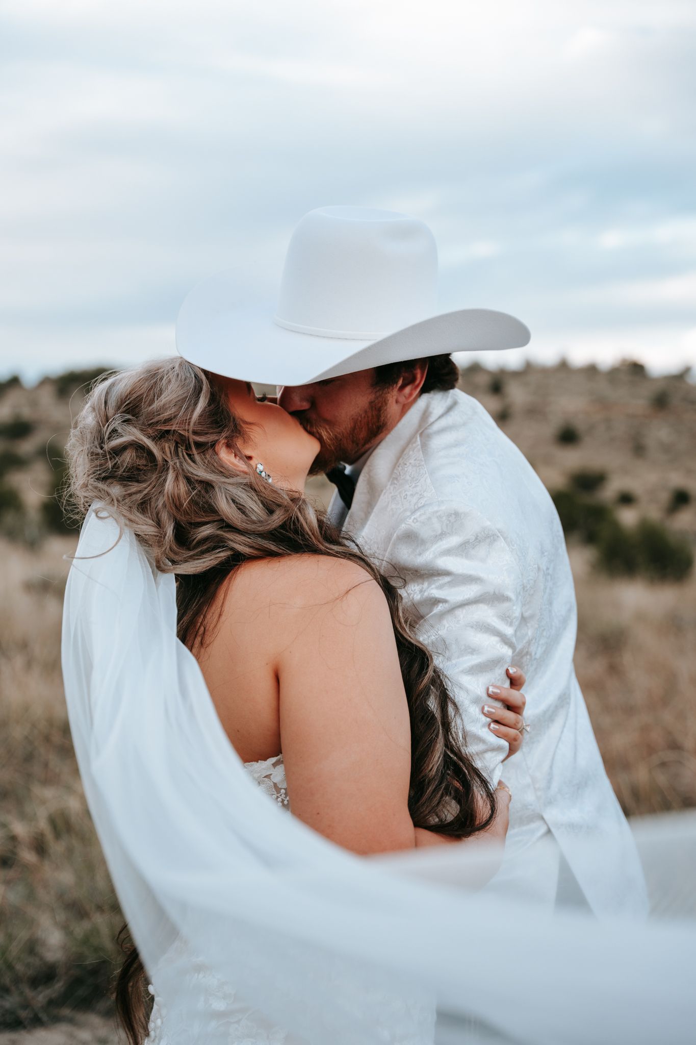 Bride and groom share passionate kiss outdoors, groom wearing white cowboy hat and jacket in rustic desert setting.