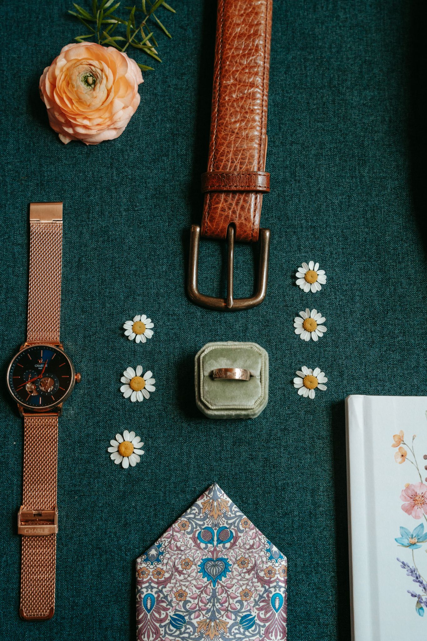 Flat lay of groom wedding accessories including rose gold watch, brown leather belt, ring, pocket square, and daisies.