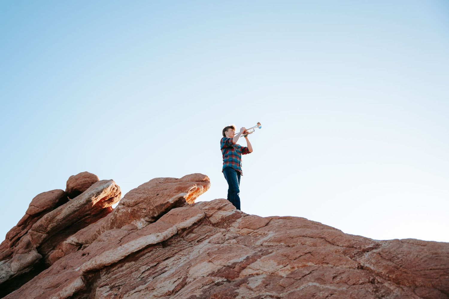 Person playing trumpet while standing atop red rock formation against clear blue sky.