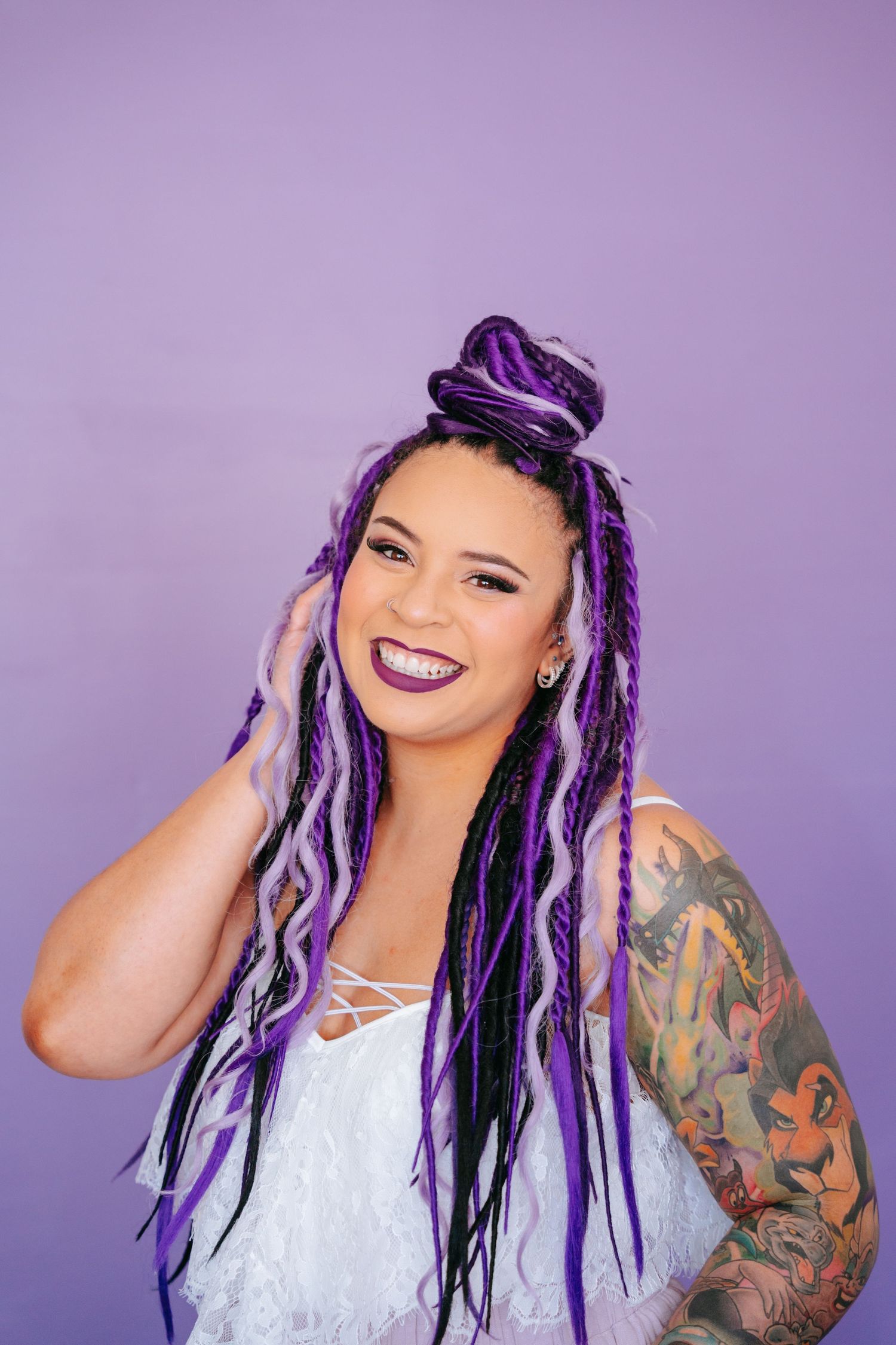 Woman with purple braids in bun, white lace top, colorful arm tattoo, laughing against a bright purple background.