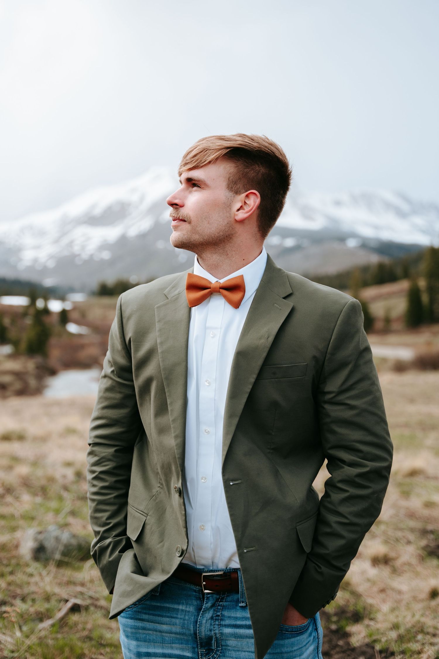 Handsome groom in olive green blazer and rust bow tie stands confidently before a snowy mountain.