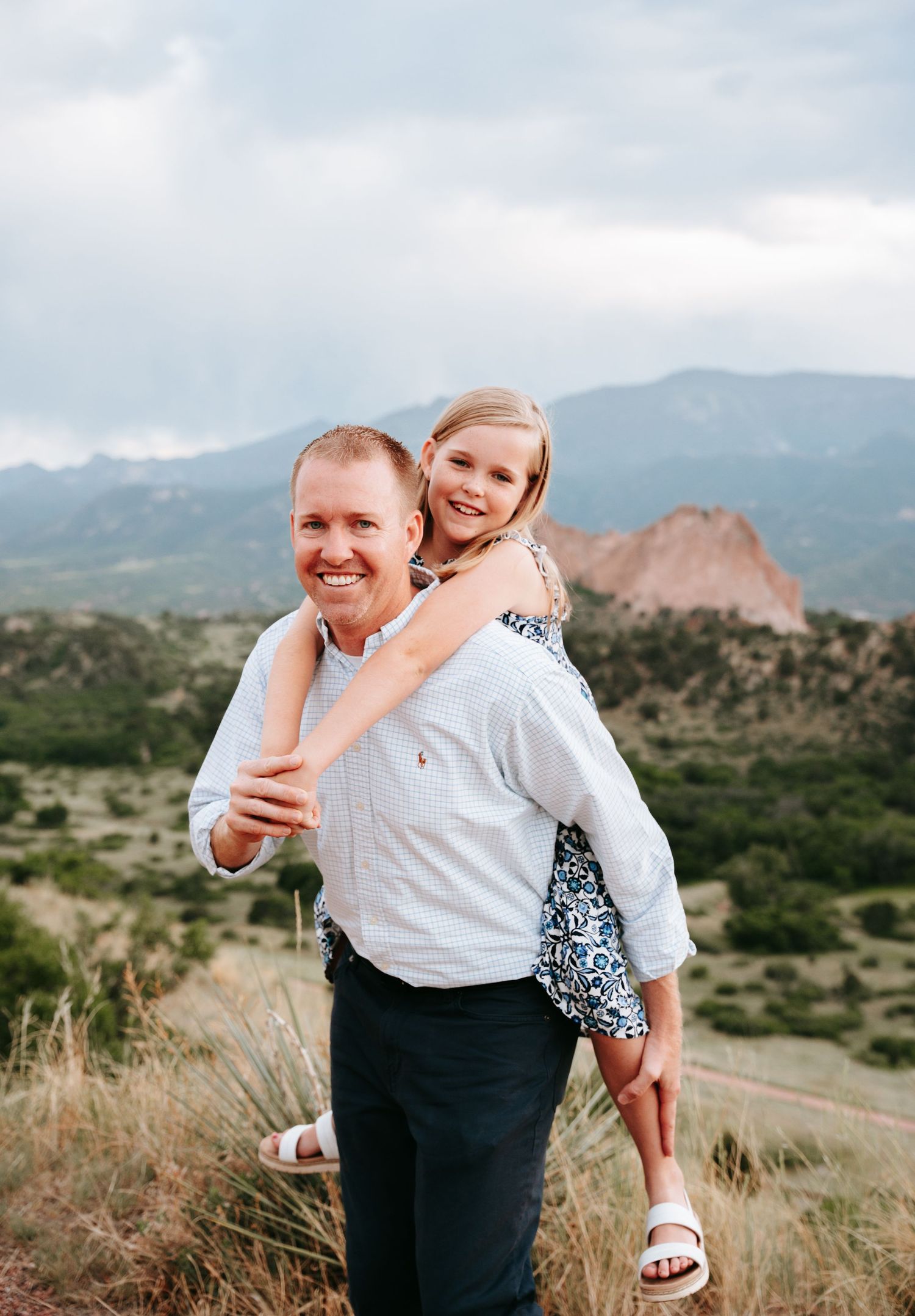Smiling man giving young blonde girl a piggyback ride with Colorado mountains in background.