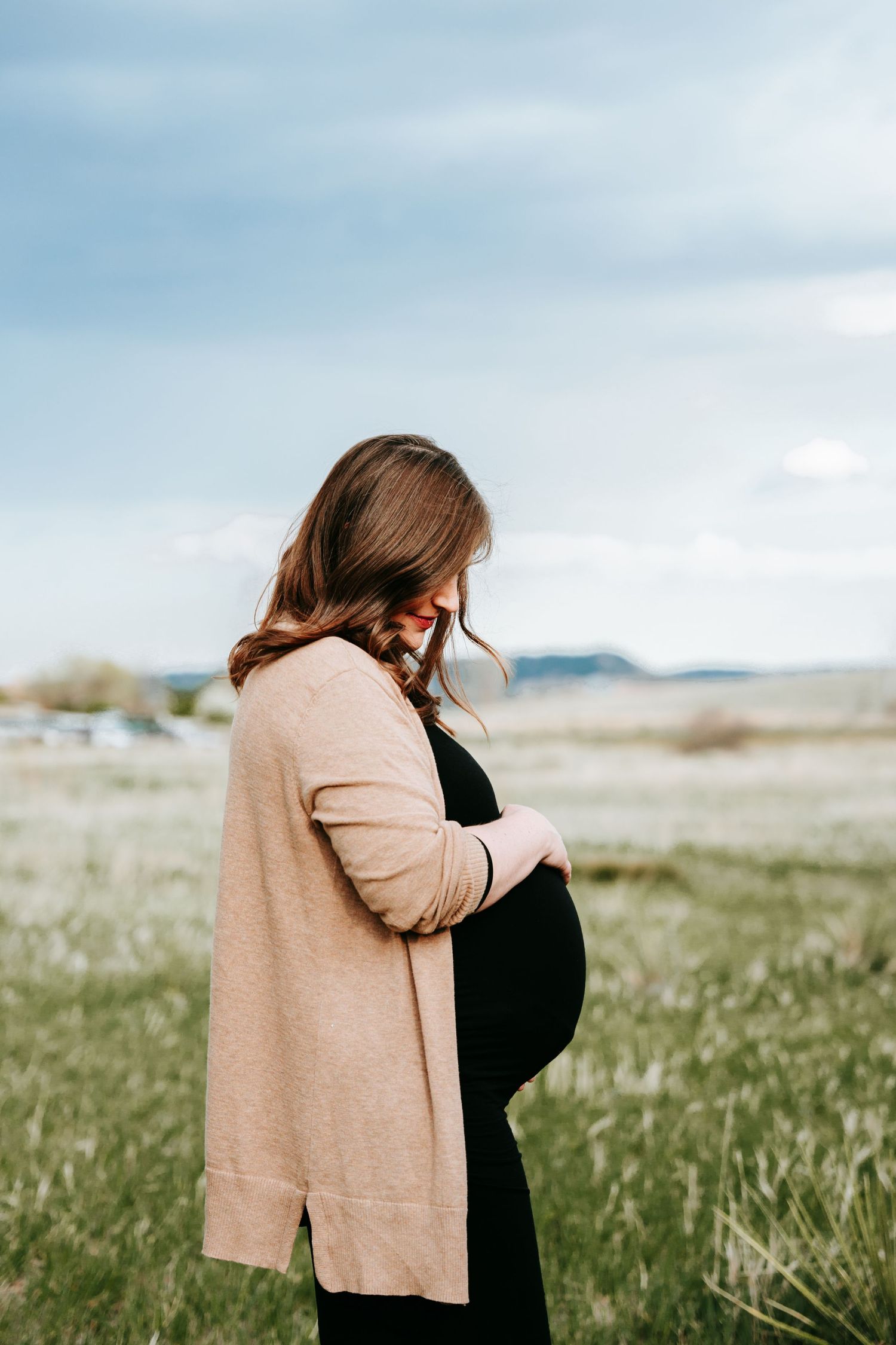 Pregnant woman in pink cardigan and black dress cradling baby bump in a peaceful open field.