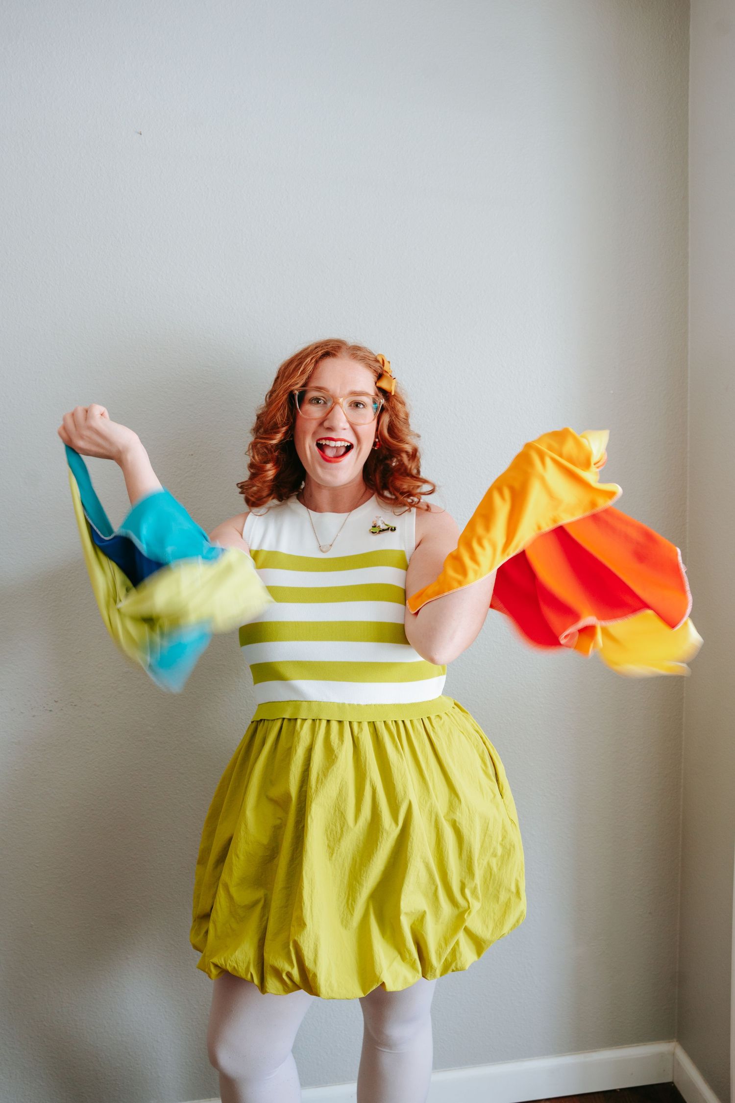 Excited redhead woman in green striped dress waving colorful scarves against white wall.