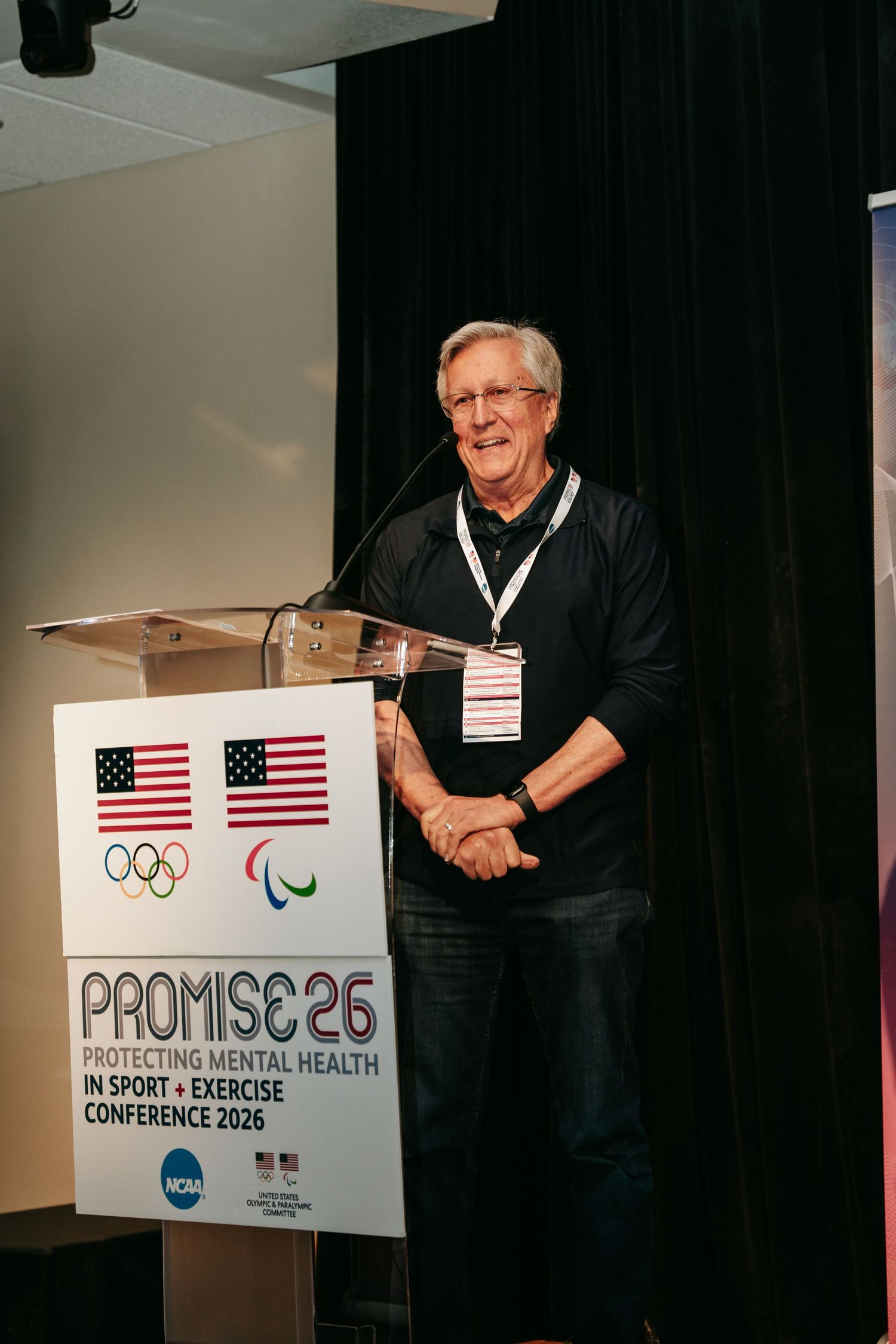 Man in glasses and a black shirt speaks at a podium at the PROMISE 26 conference on protecting mental health in sport and exercise.