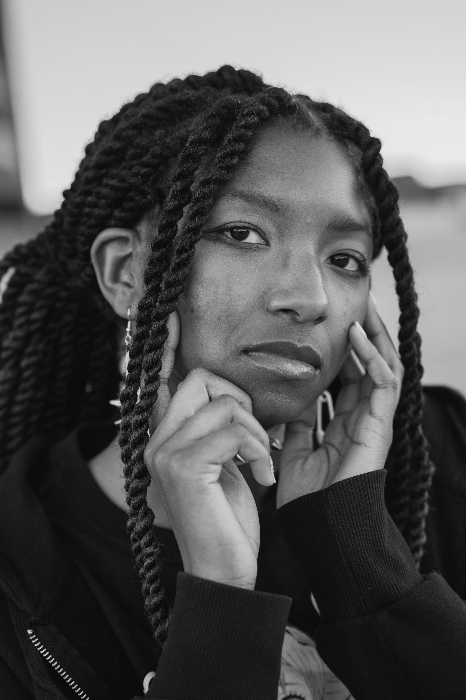 Black and white portrait of a young woman with braided hair posing thoughtfully with hands near face.