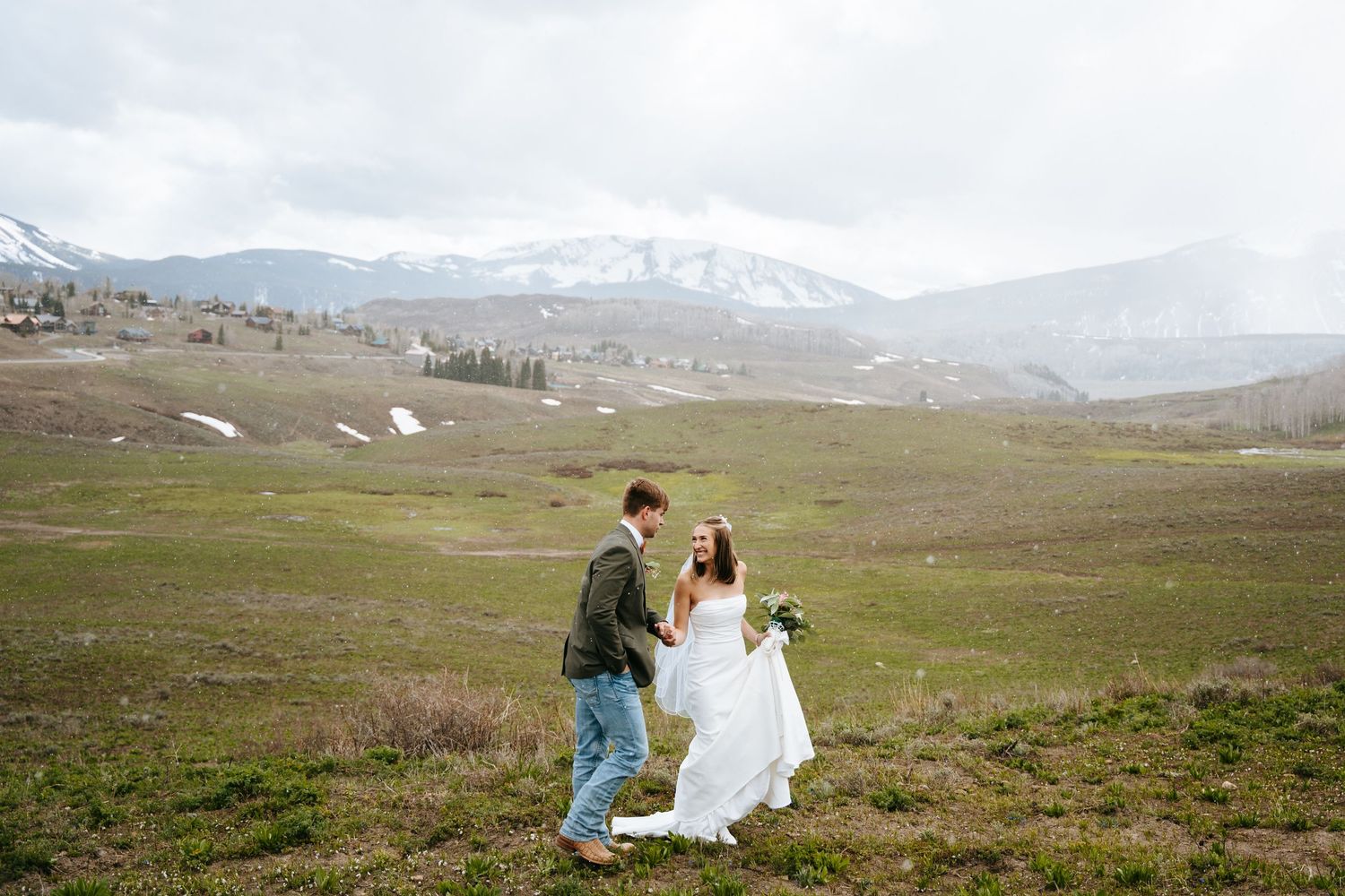 A couple in wedding attire stands in a scenic mountain meadow in Crested Butte, Colorado with snow-capped peaks in the distance.