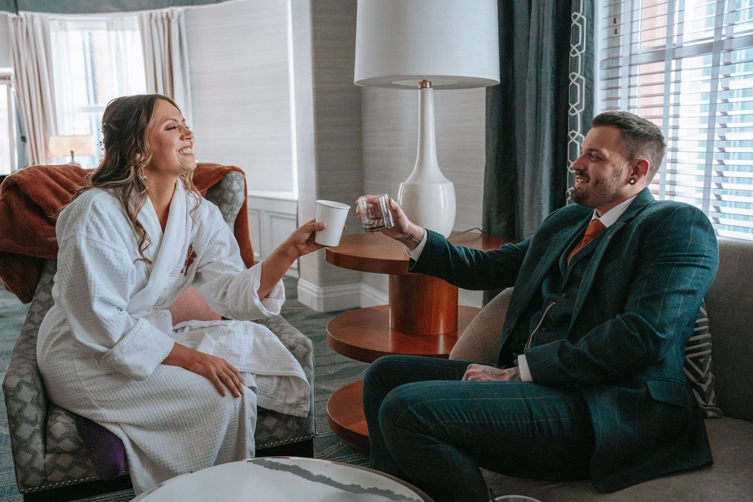 Bride in white robe and groom in teal plaid suit toast with mugs in a hotel suite.