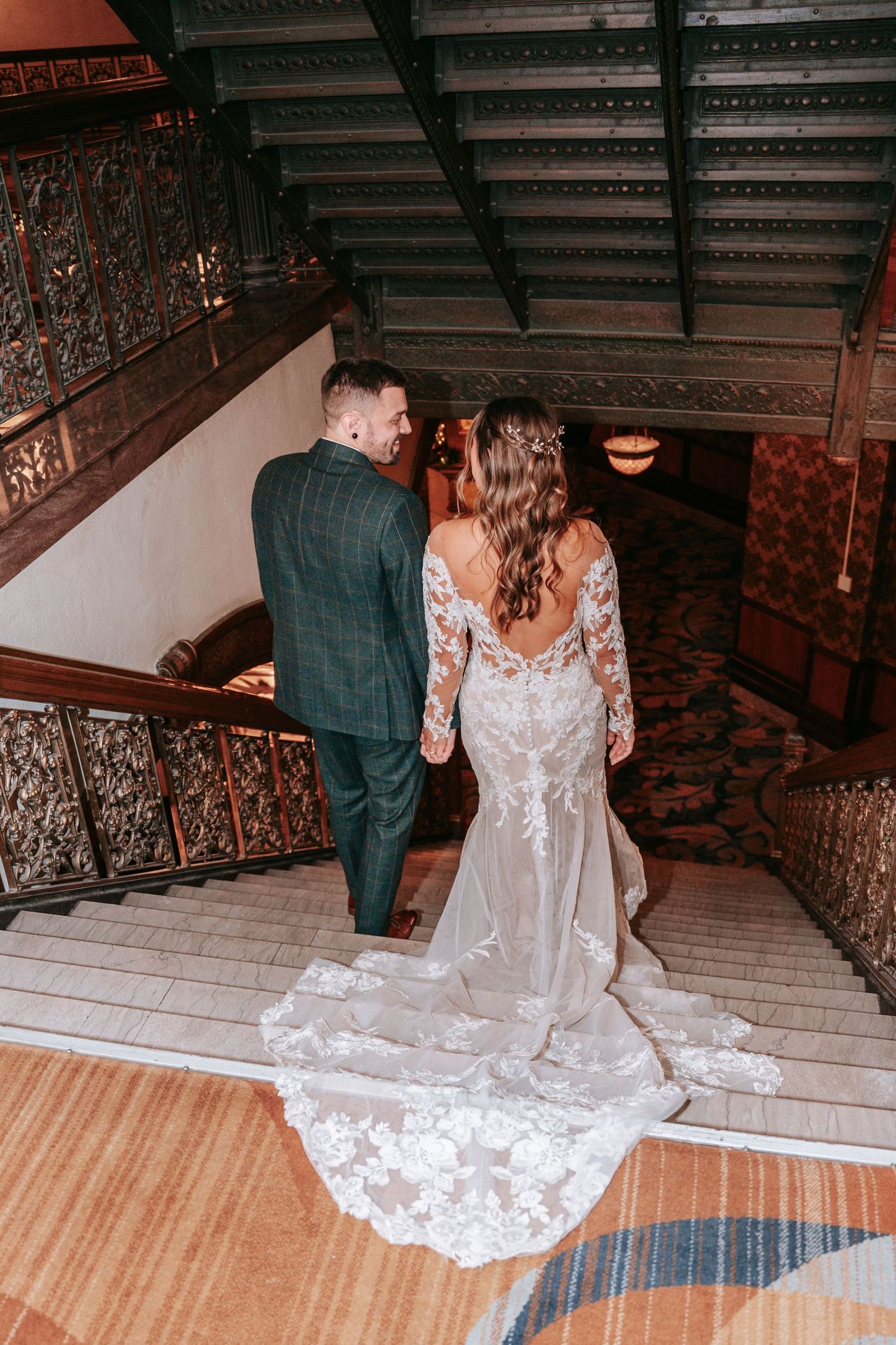 Bride and groom walk up ornate wooden staircase, bride's lace gown train trailing behind her.