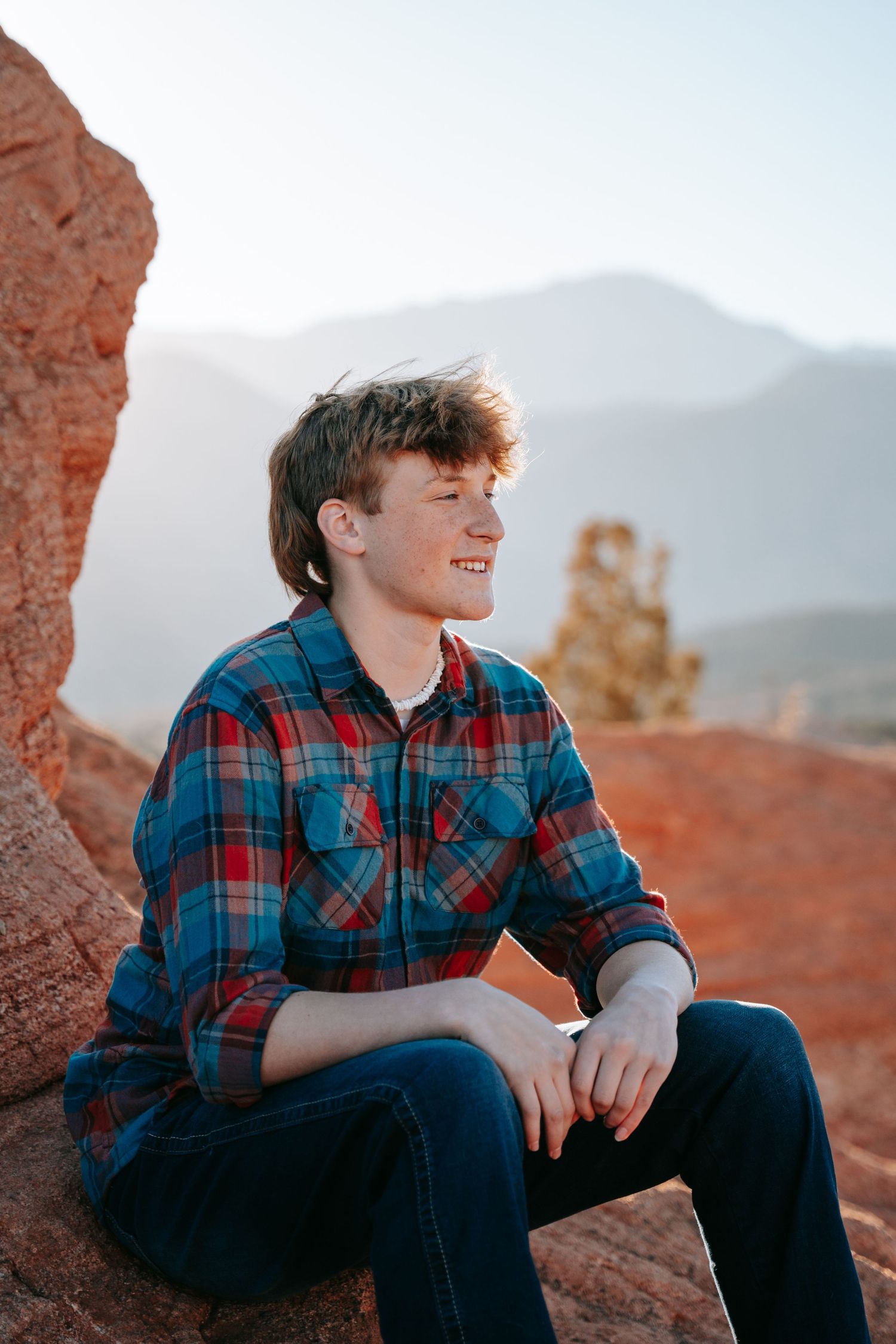Smiling teenage boy in plaid flannel shirt sitting on red rocks with mountain scenery behind him.