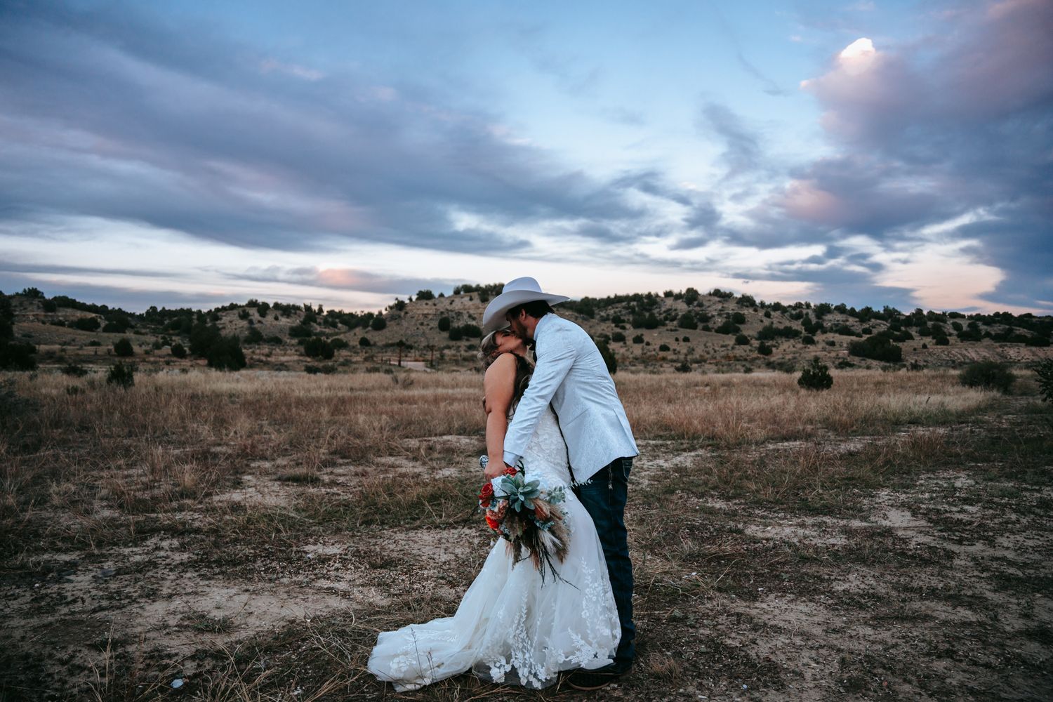 Bride and groom share a romantic kiss in an open field at sunset with dramatic cloudy skies.