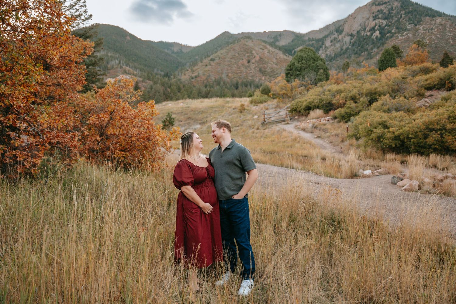 Pregnant couple standing in autumn mountain meadow, woman in red dress, man in green shirt, colorful fall foliage backdrop.