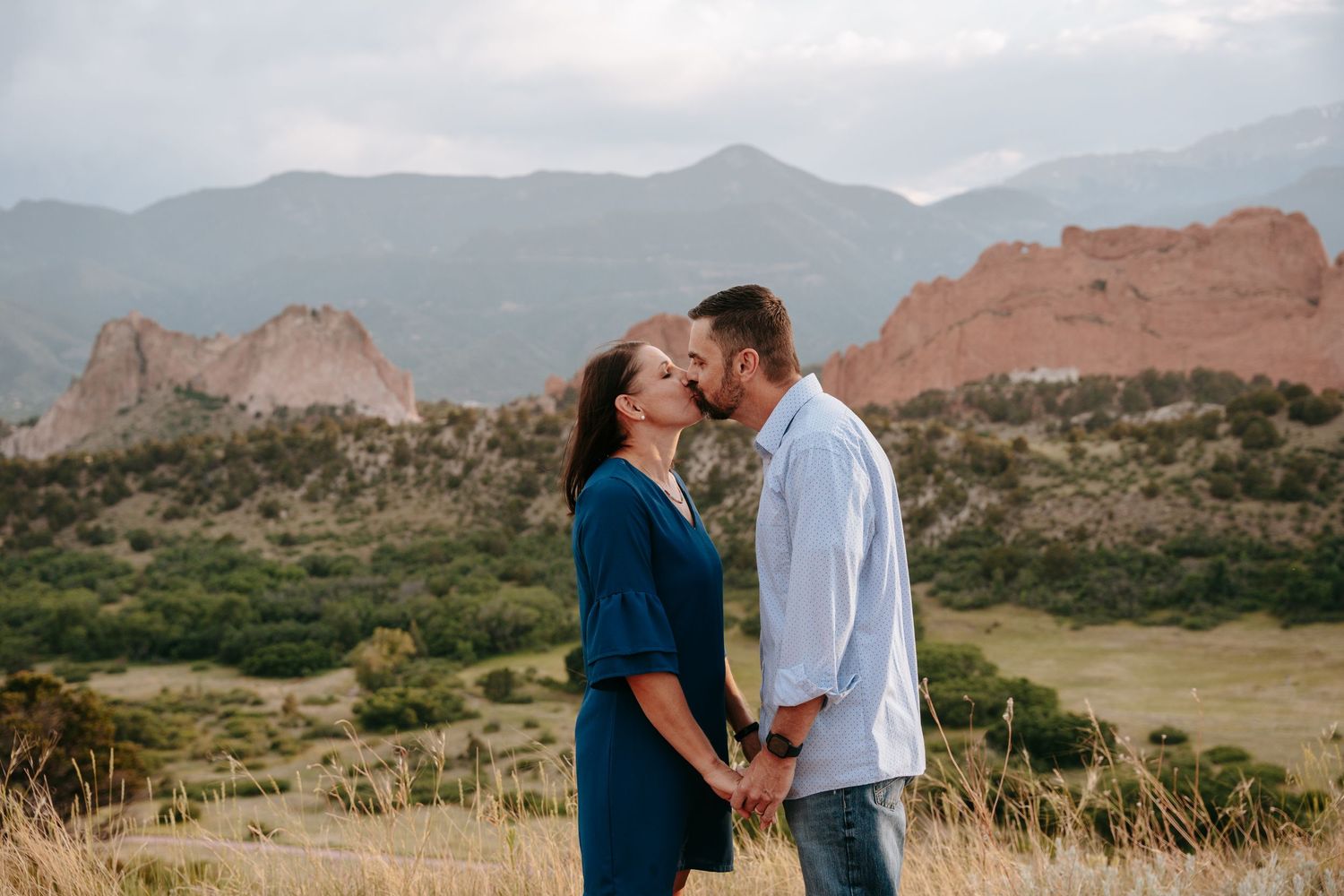 Couple sharing a kiss while holding hands in a mountain meadow with red rock formations.