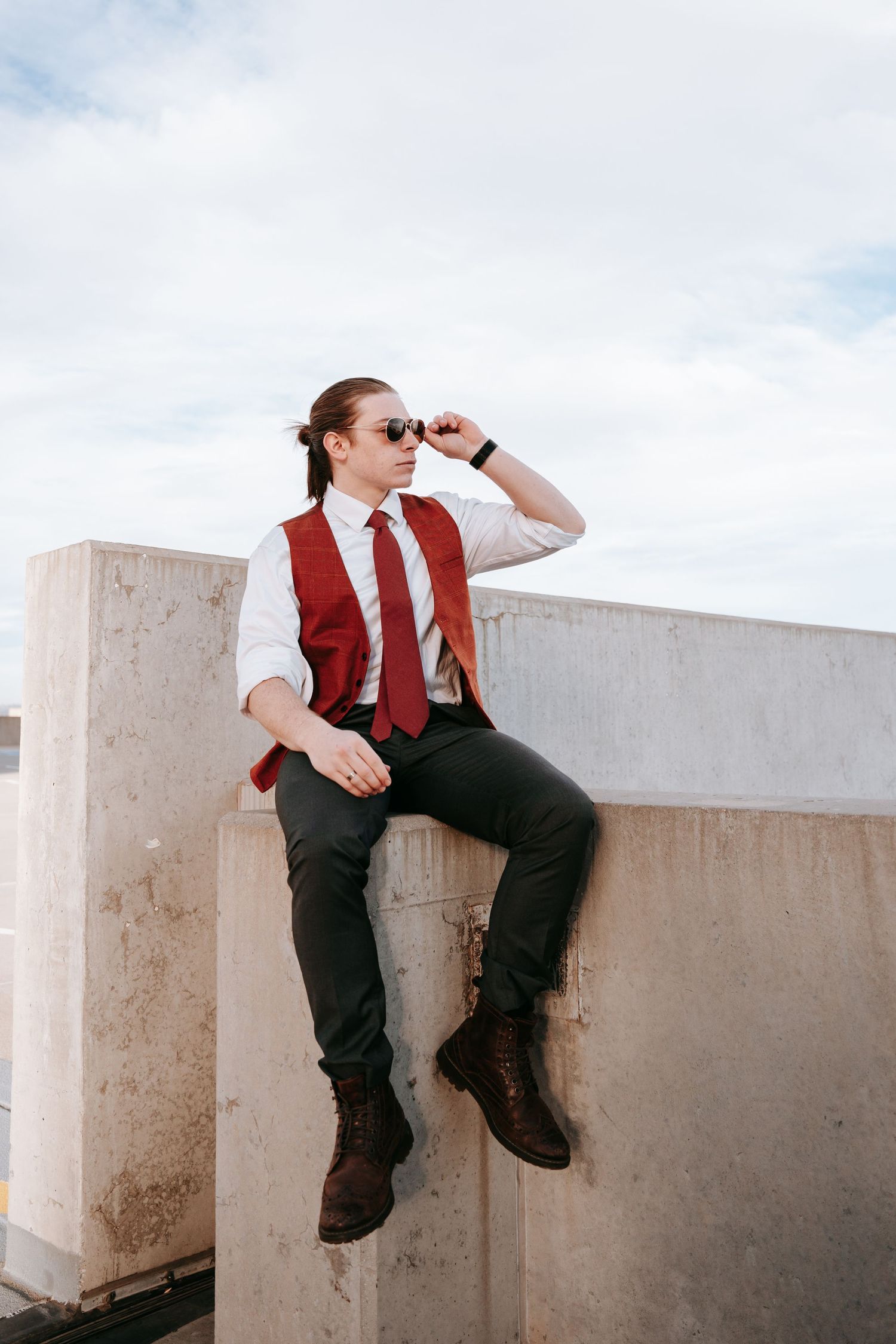 A stylish young man in a red vest and tie sitting on a concrete wall outdoors.