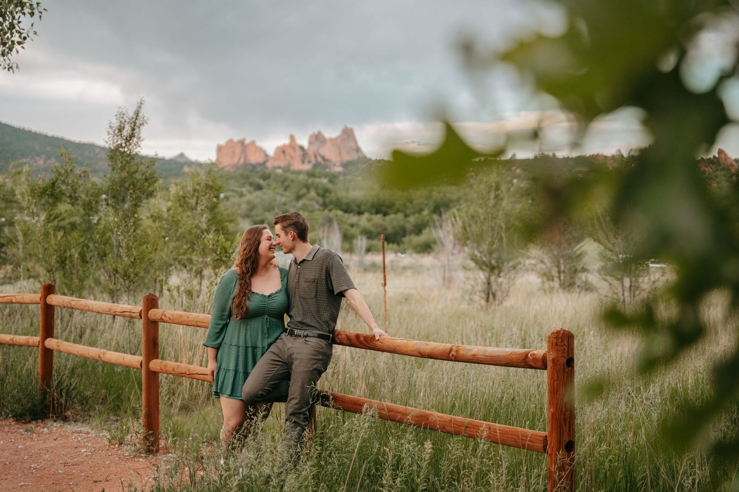 Couple kissing by a wooden fence with red rock formations and lush greenery at Garden of the Gods, Colorado.