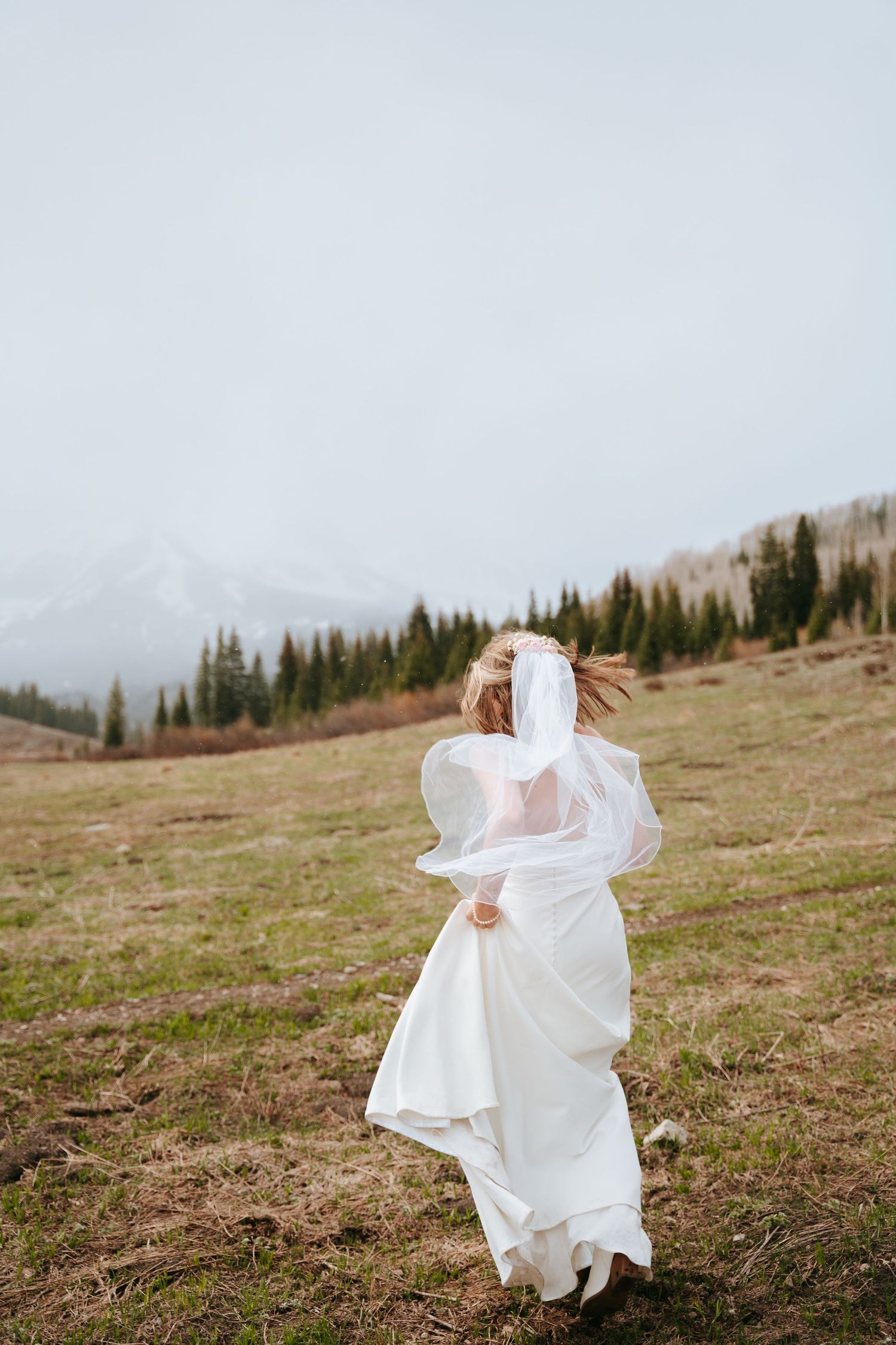 Bride in white wedding dress and veil running through mountain meadow on cloudy day.