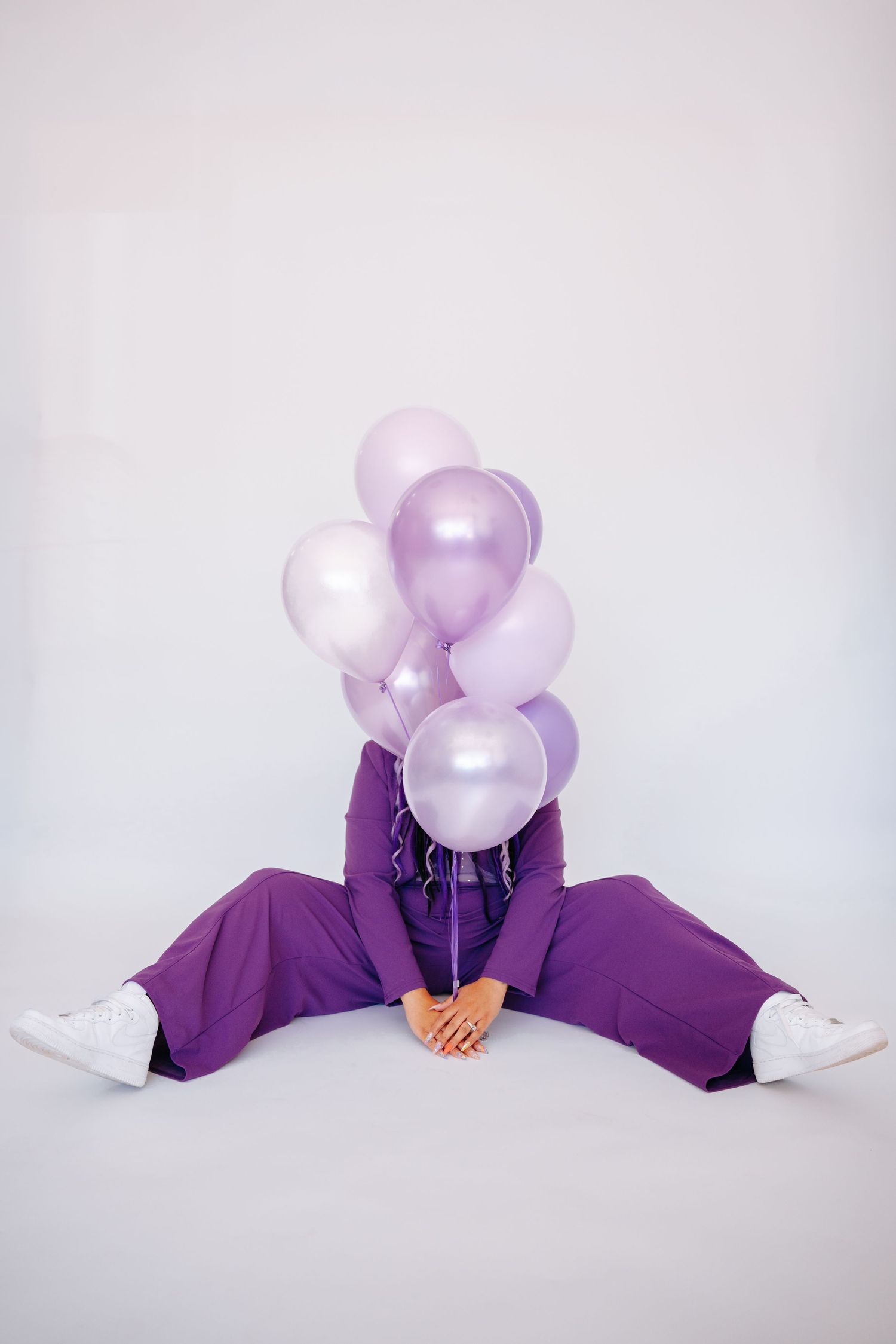 Person in purple suit sitting on white floor with face hidden behind cluster of lavender and pearl balloons.