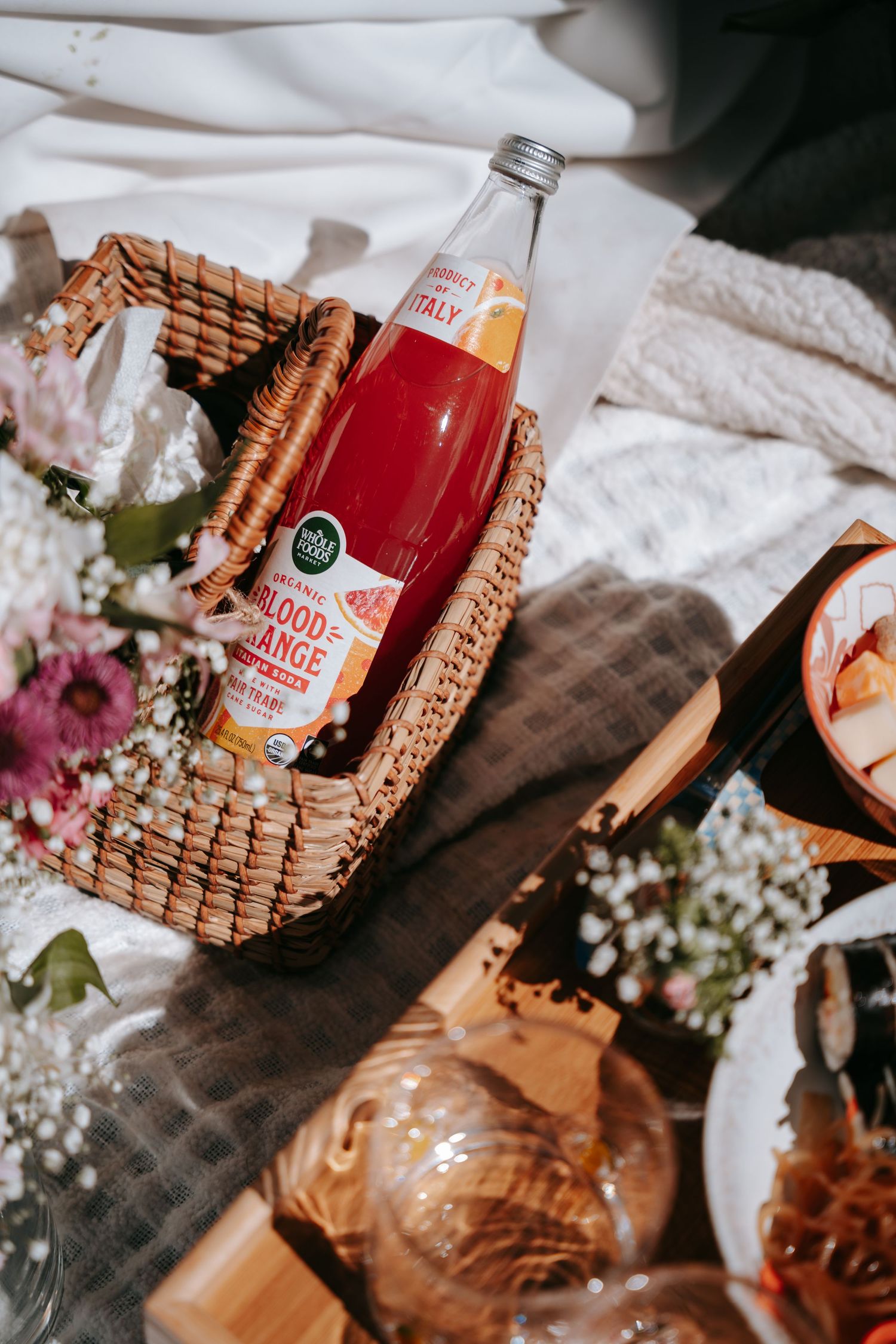 Wicker picnic basket with blood orange Italian soda bottle, surrounded by fresh flowers on a white blanket.