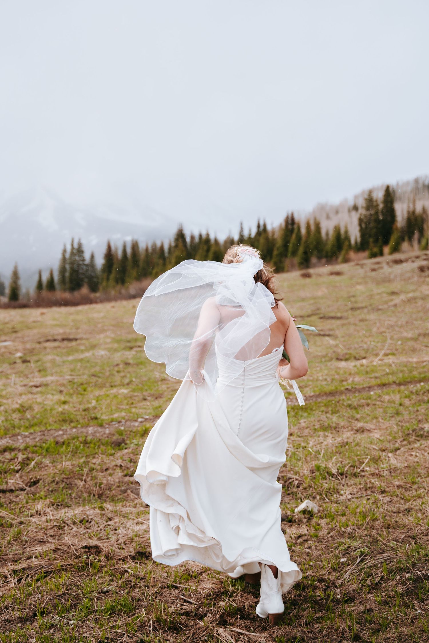 Bride in white gown and boots with veil blowing in wind on mountain meadow.