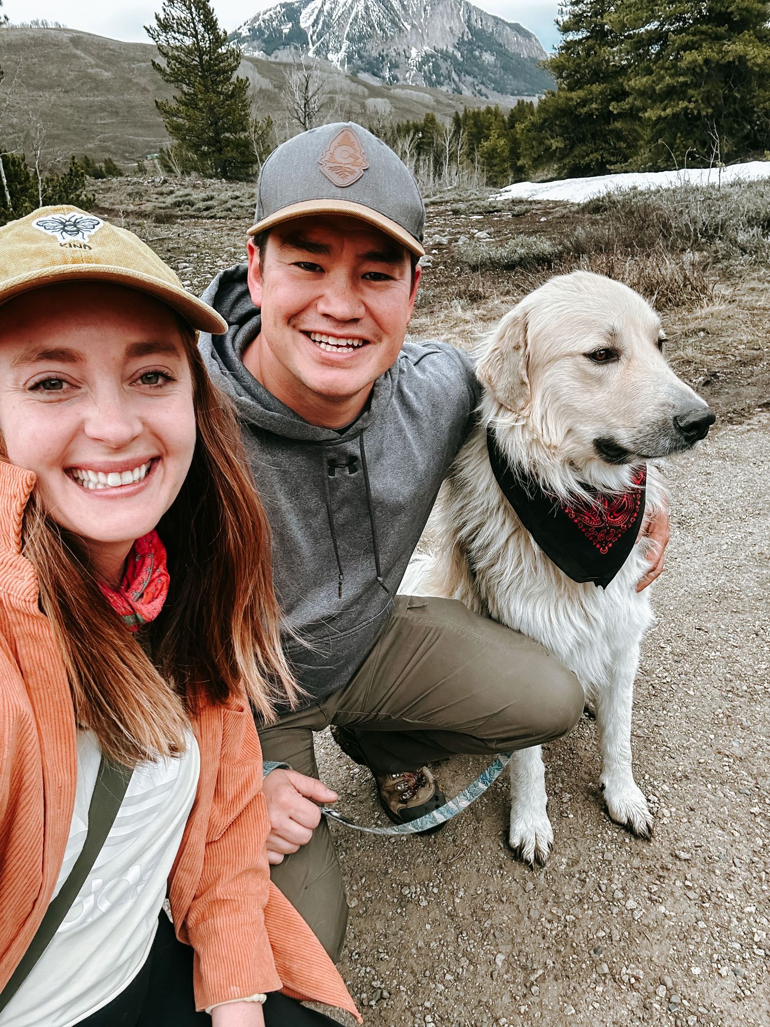 Smiling couple posing with white golden retriever wearing red bandana on mountain trail.