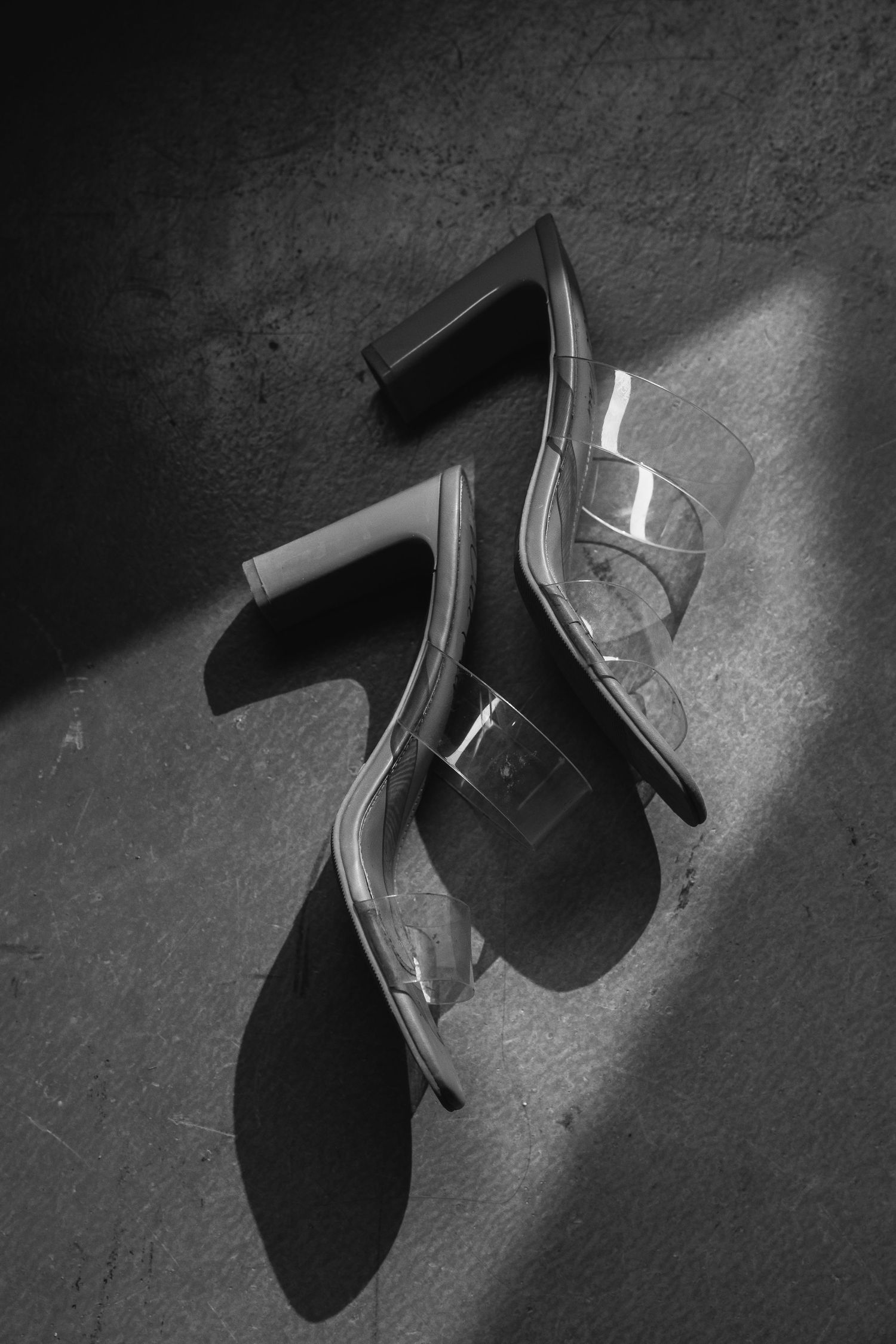 Black and white photo of clear perspex block heel mules on textured surface with dramatic shadow lighting.