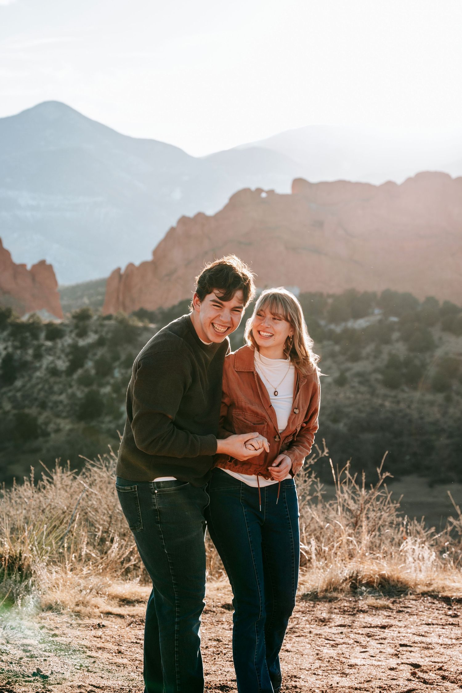 Happy couple holding hands smiling at Garden of the Gods Colorado with sunlit red rock formations behind them.