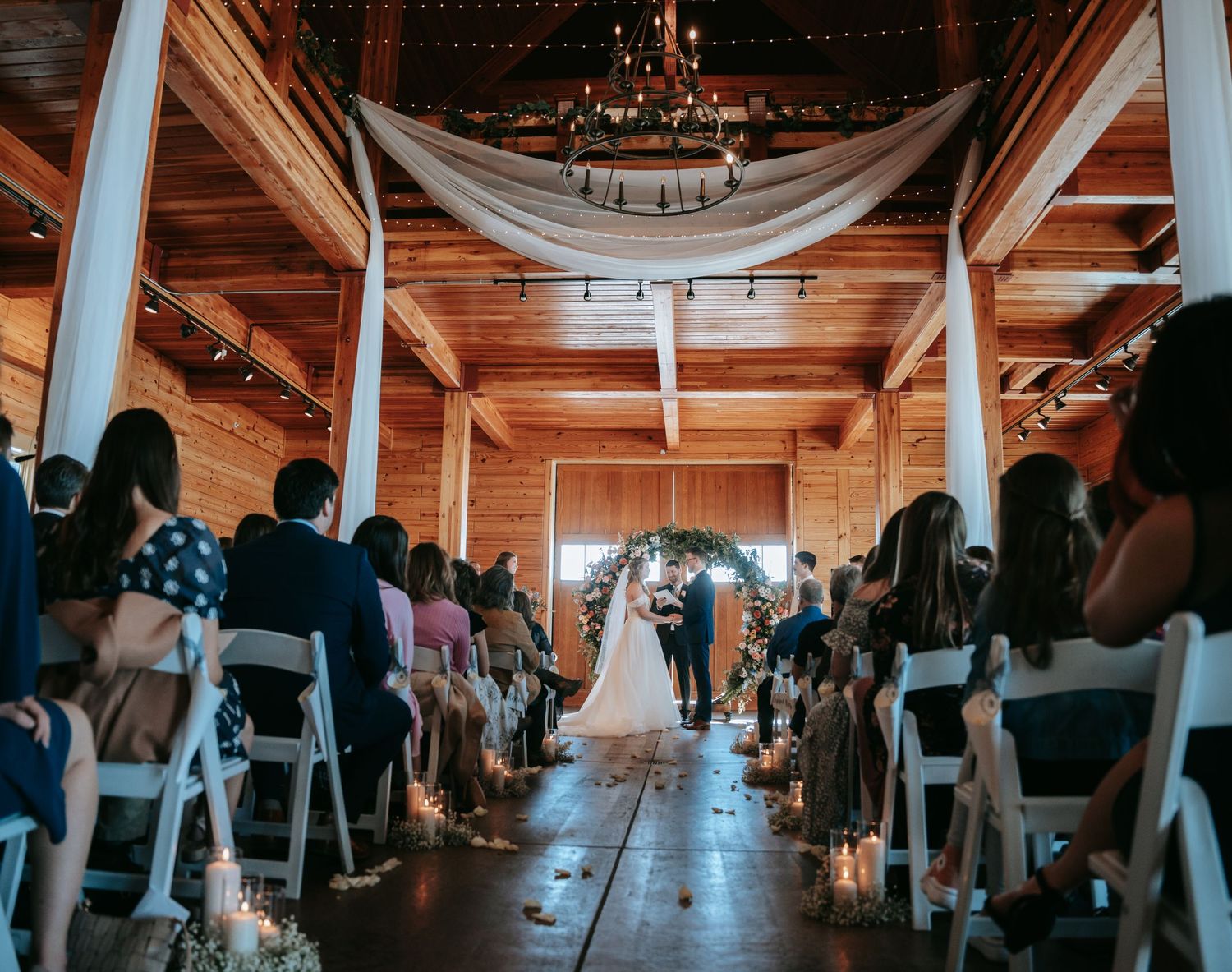 Wedding ceremony in rustic barn venue with floral arch and white chairs.