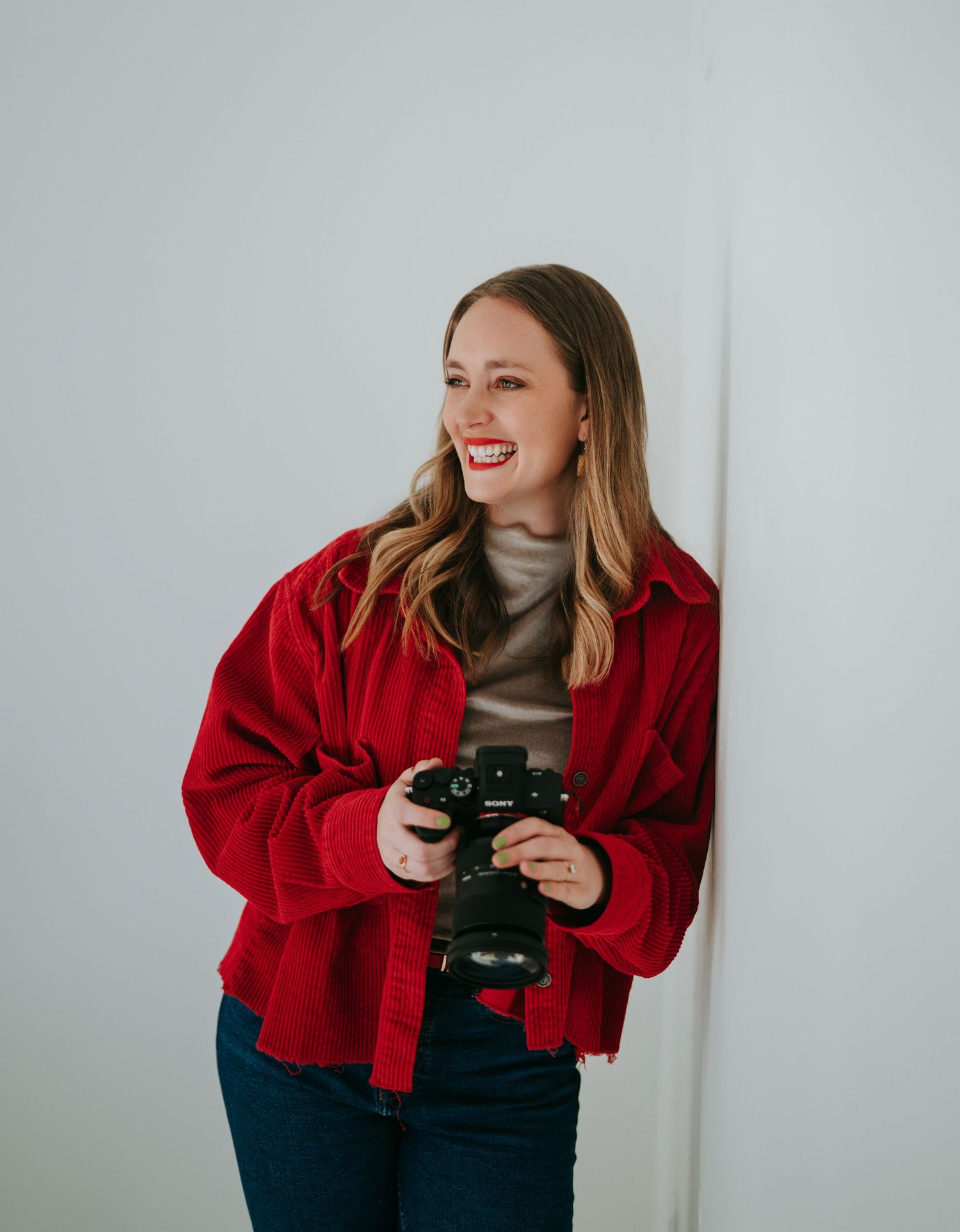 Smiling female photographer in red jacket holding camera against white wall.