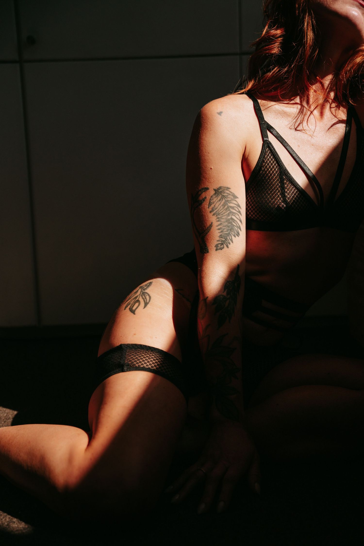 Woman in black lingerie with floral tattoos on arm and thigh, posed in dramatic shadow lighting.
