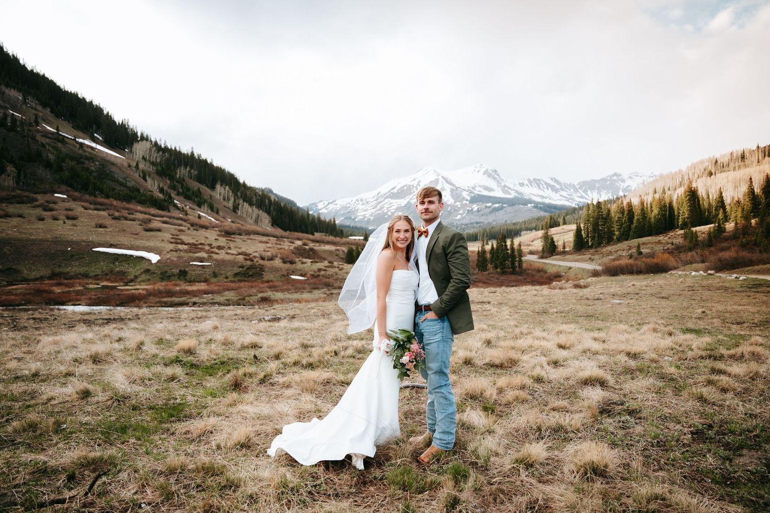 Smiling bride and groom pose together in a dry mountain meadow surrounded by snow-capped peaks.