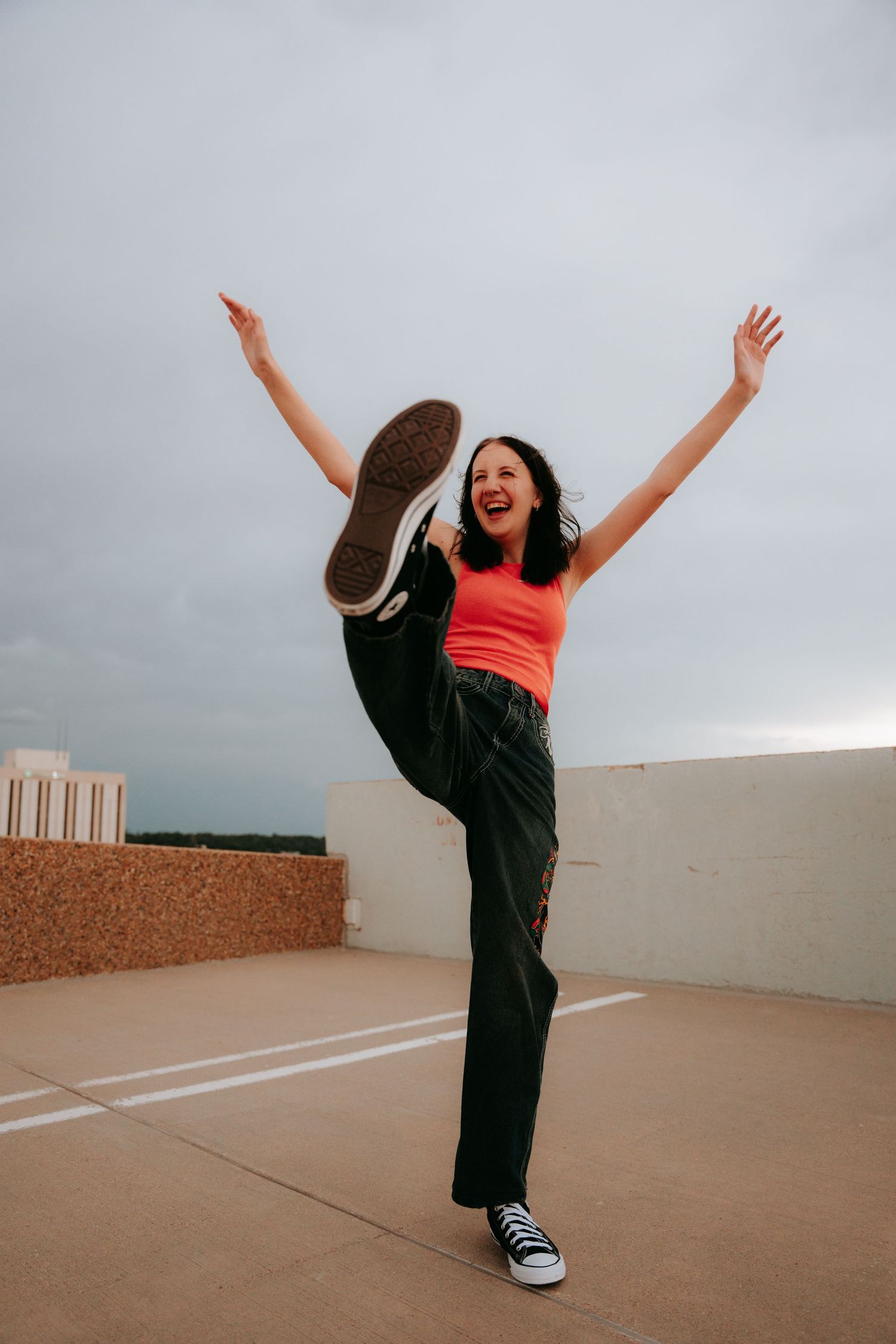 Energetic young woman in orange top jumping with leg raised on rooftop.