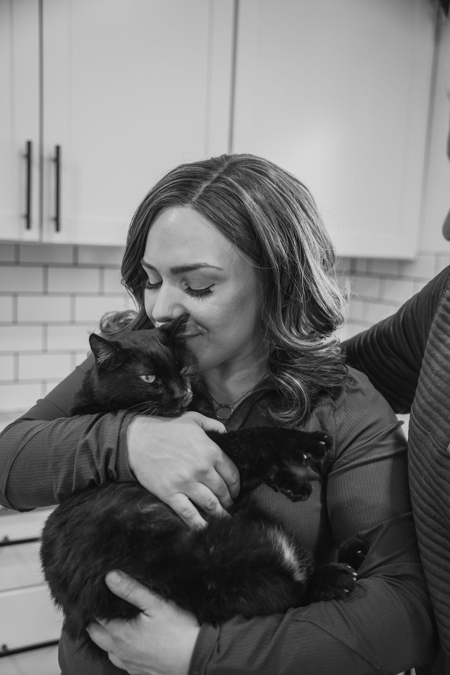 Black and white photo of a woman lovingly nuzzling a black cat in a modern kitchen.