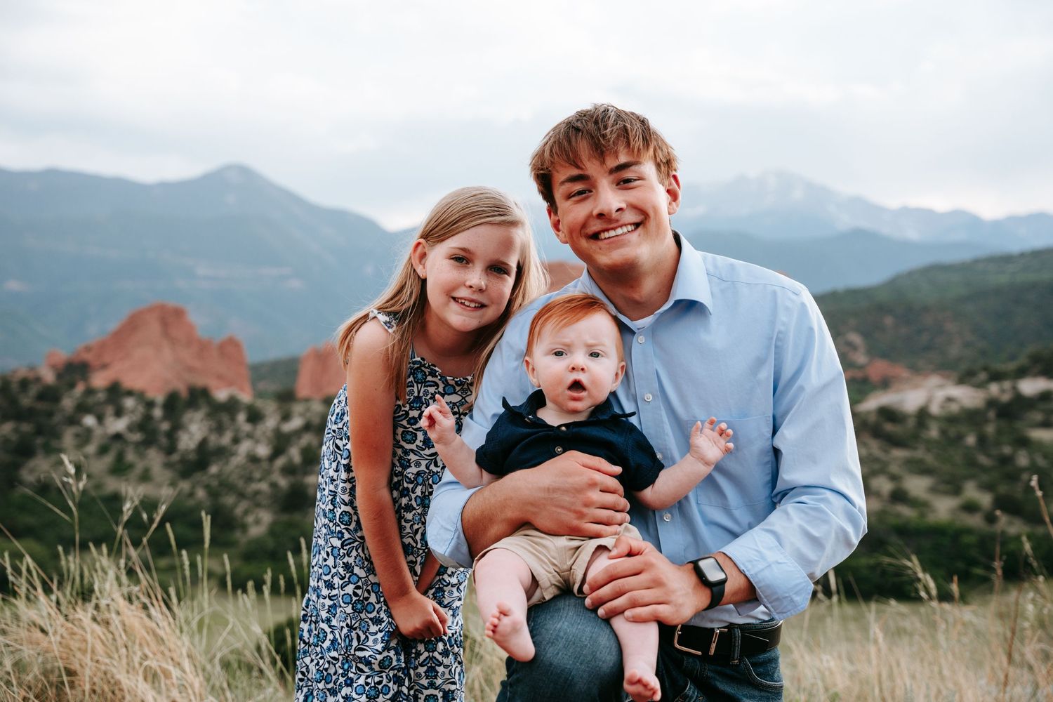 Young man smiling with two children in front of red rock formations and mountains in Colorado.