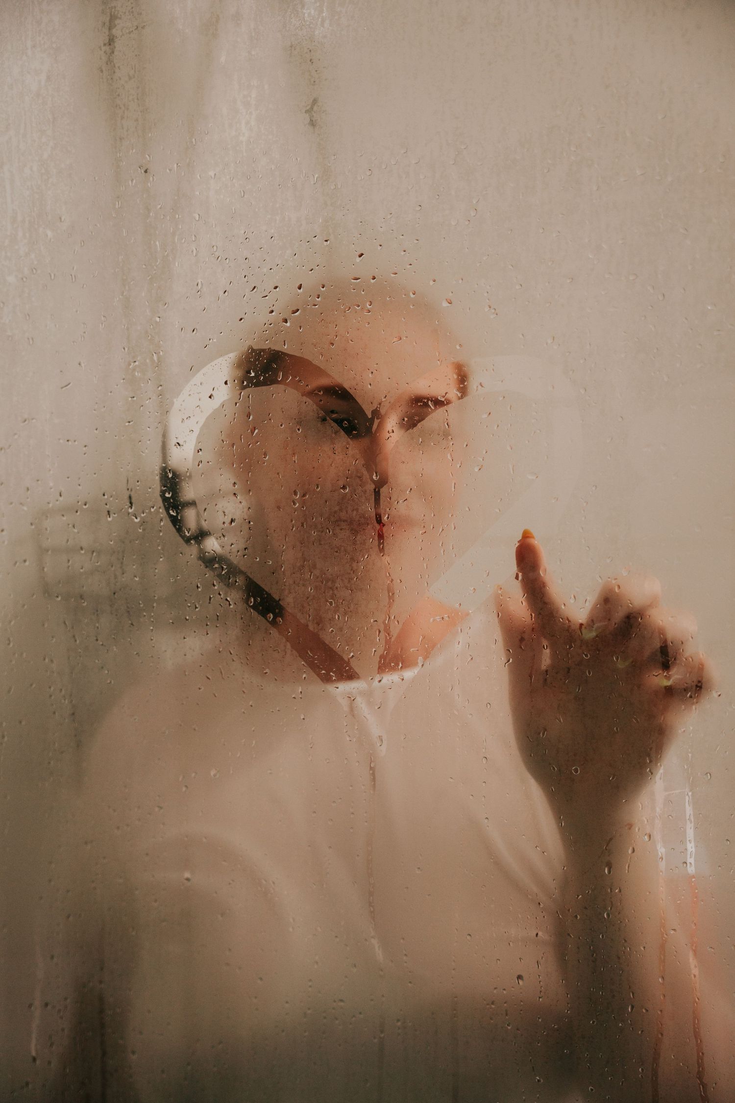 Person behind foggy wet glass with water droplets, reaching hand forward in moody atmospheric portrait.