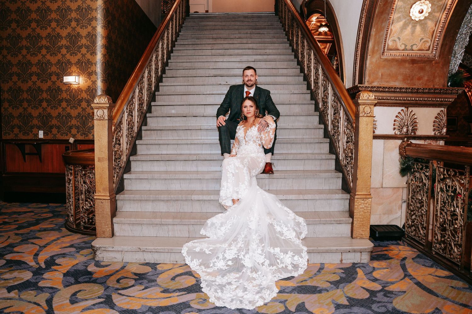 Wedding couple poses on grand staircase, bride in long lace train gown, groom in dark green suit.