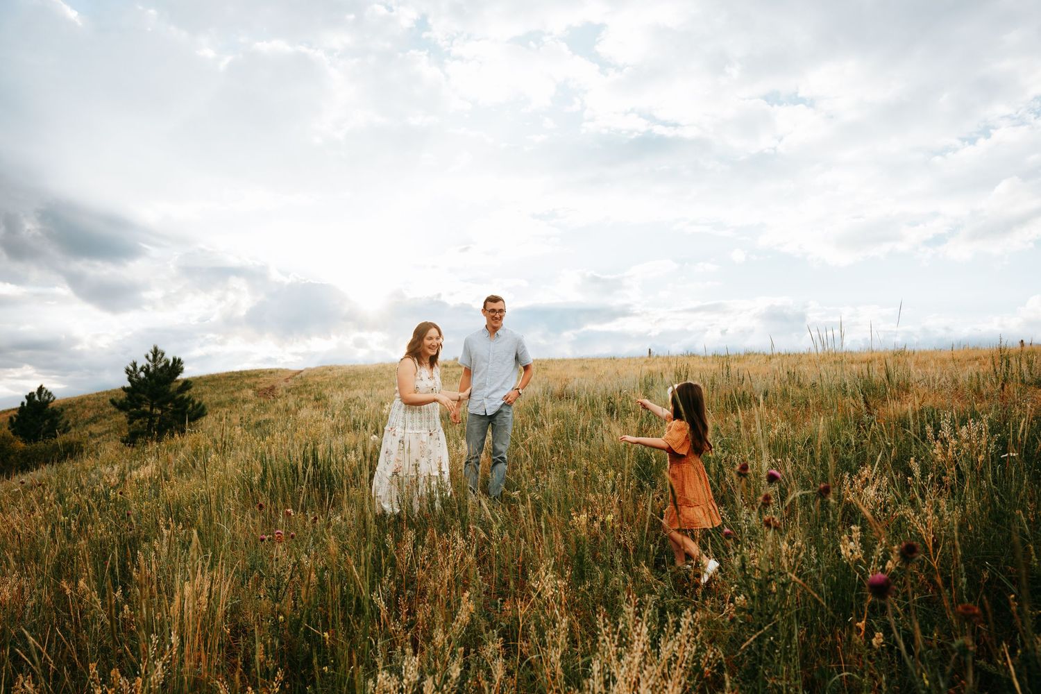 Happy family of three walking through golden meadow under cloudy sky in casual outdoor portrait.