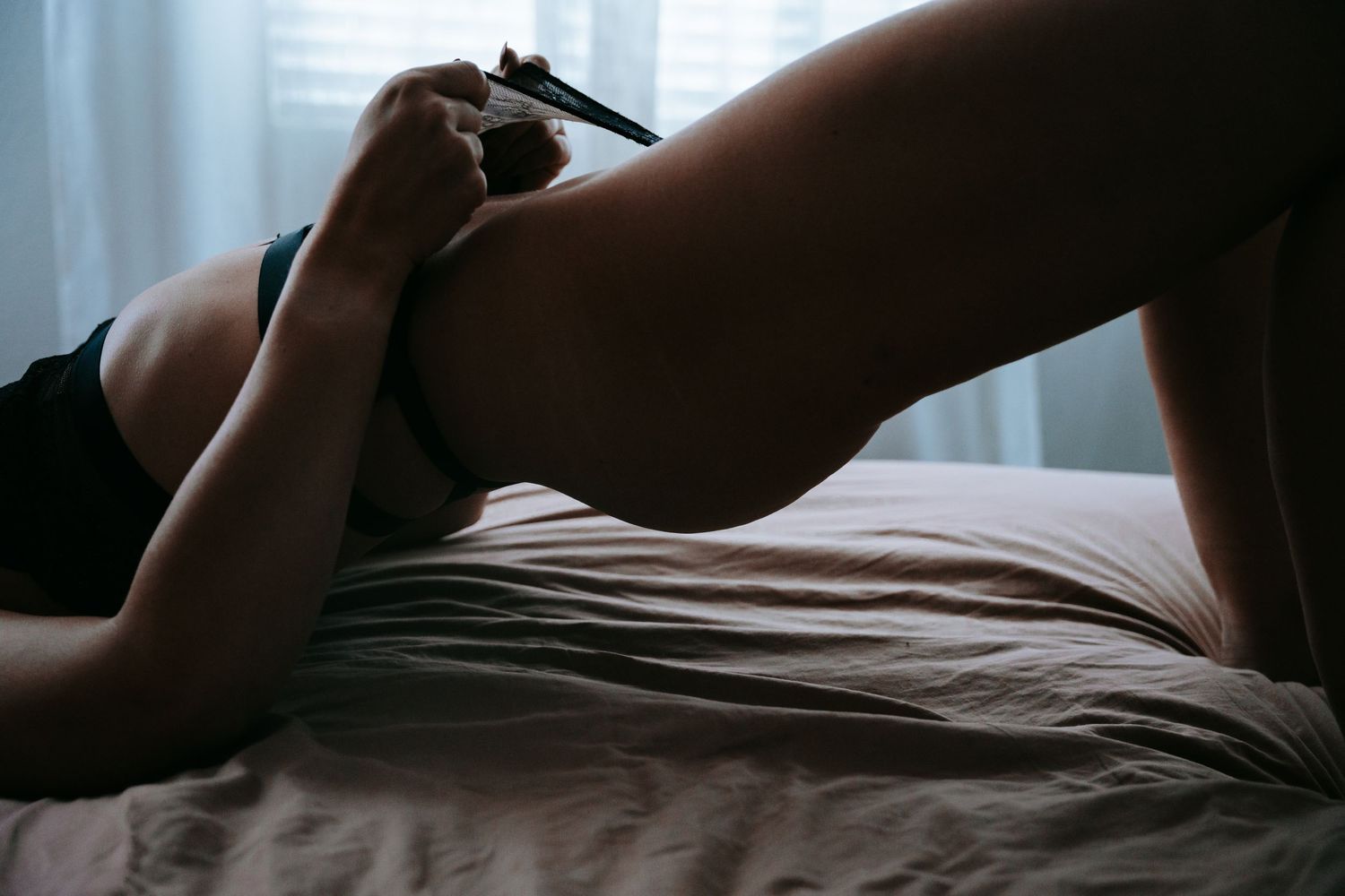 Woman in black lingerie lying on a bed in a dimly lit bedroom.