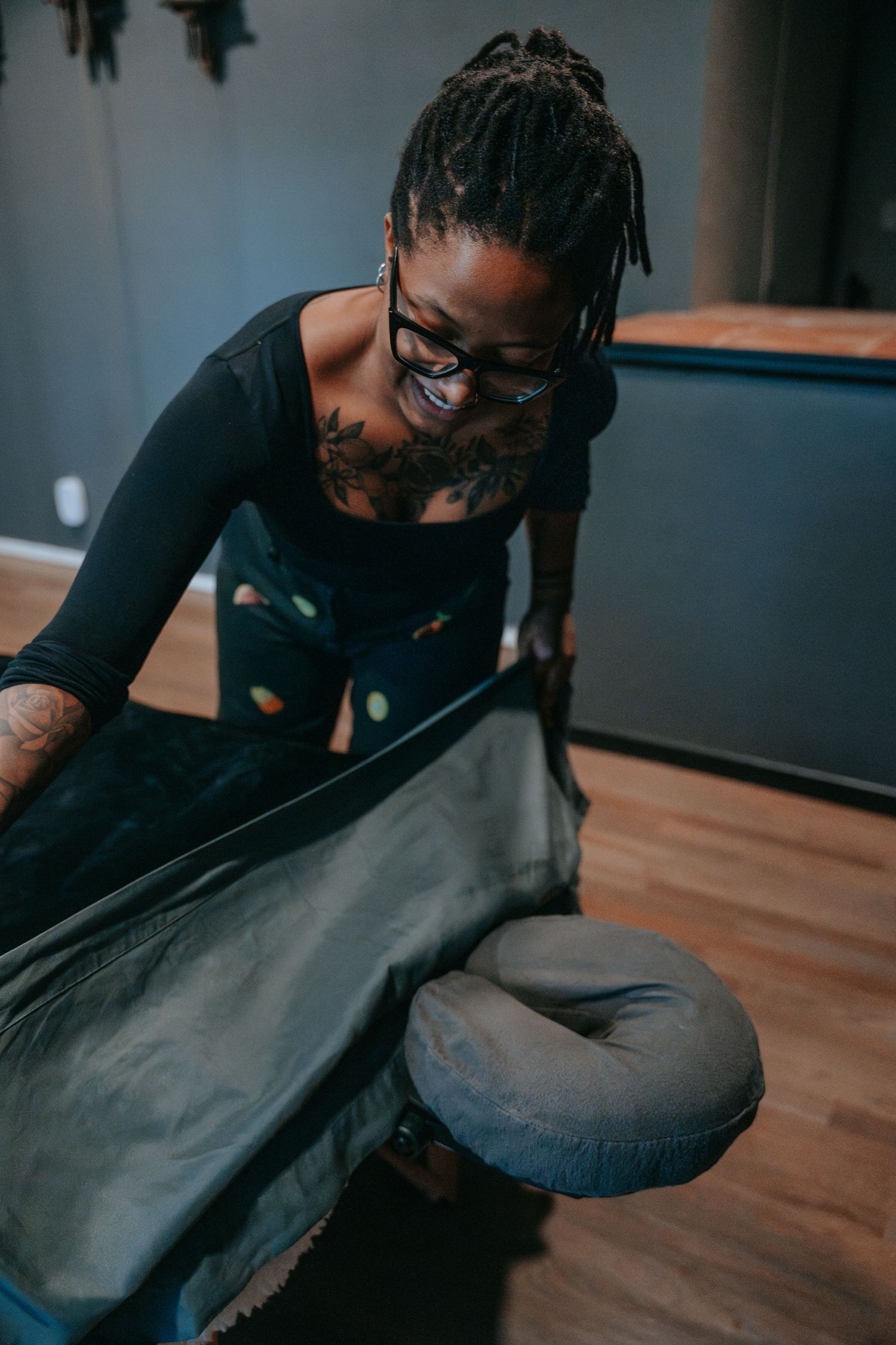 Tattooed female massage therapist with dreadlocks preparing a massage table by adjusting the sheet and face cradle.