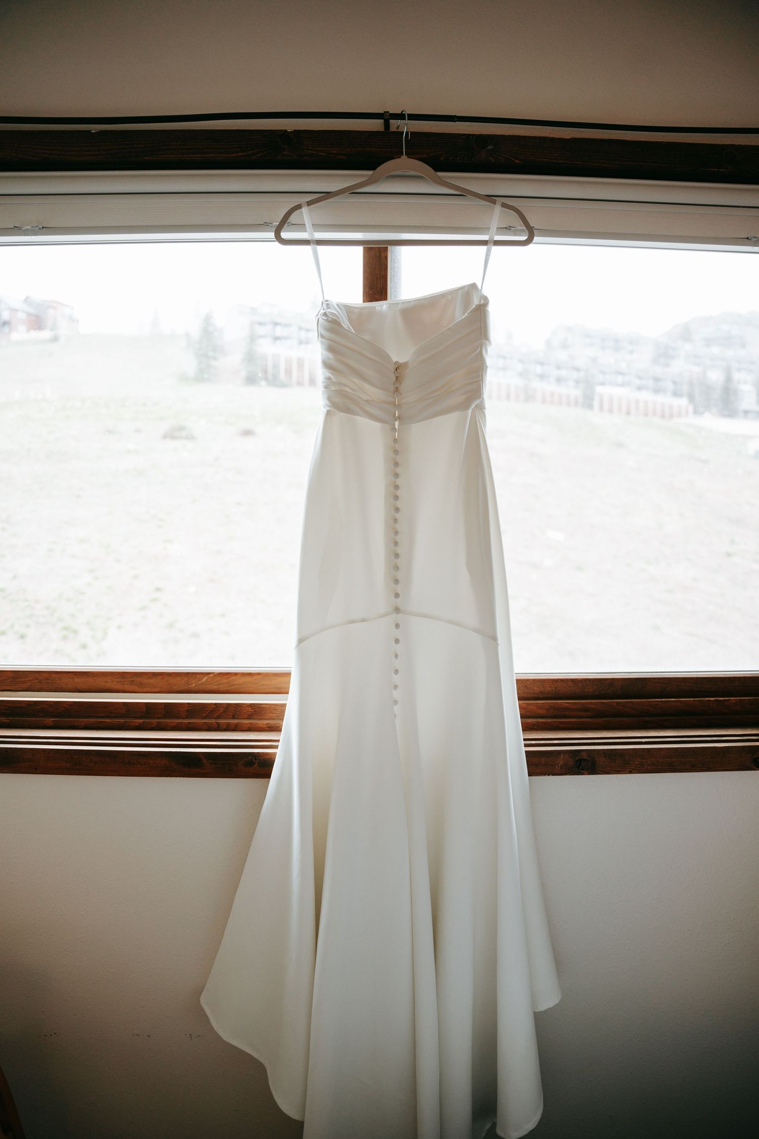 Elegant white strapless wedding gown with button back detail hanging in a window with scenic view.