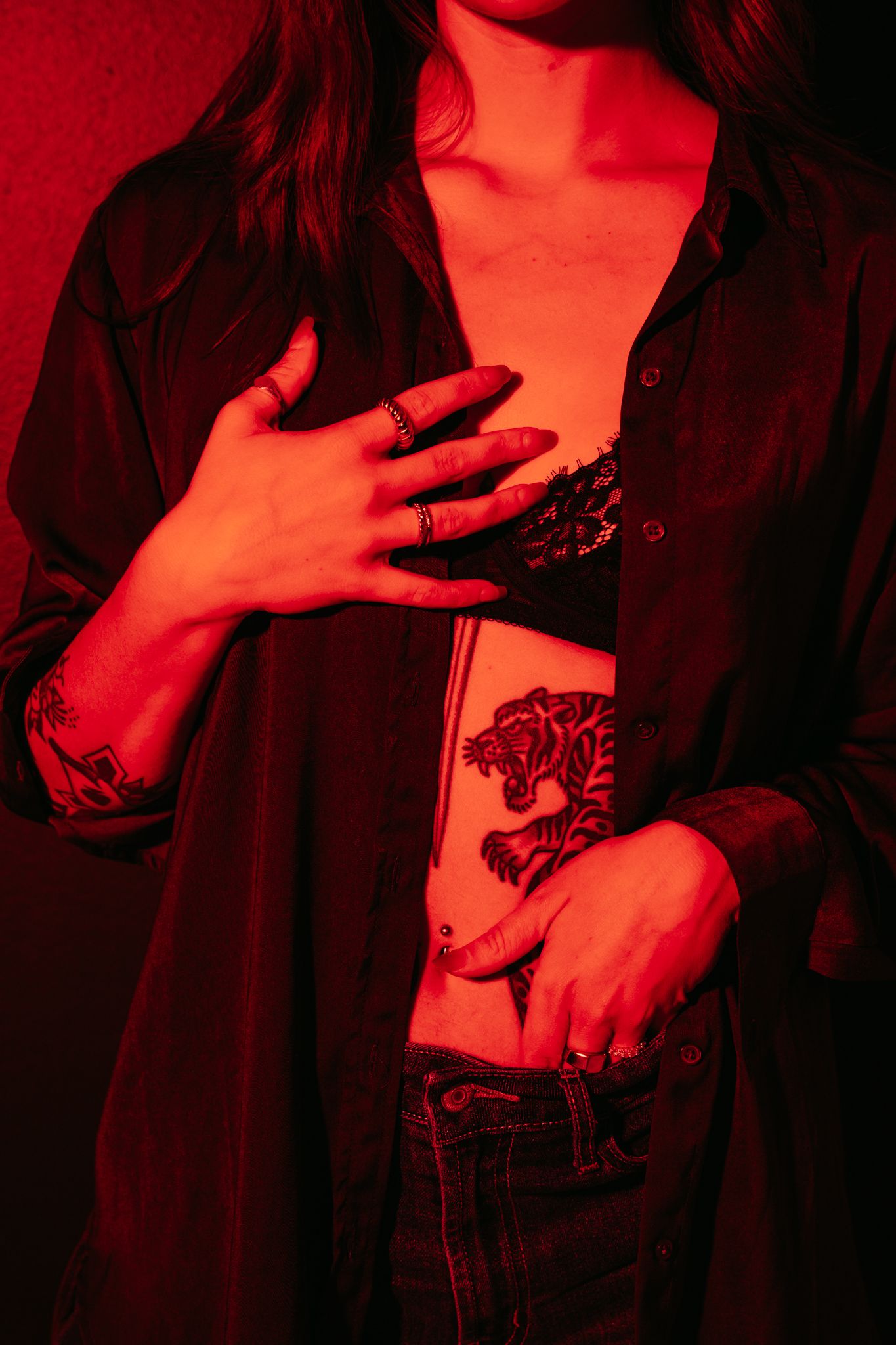 Woman in open black shirt revealing lace bralette and tiger tattoo under red lighting.