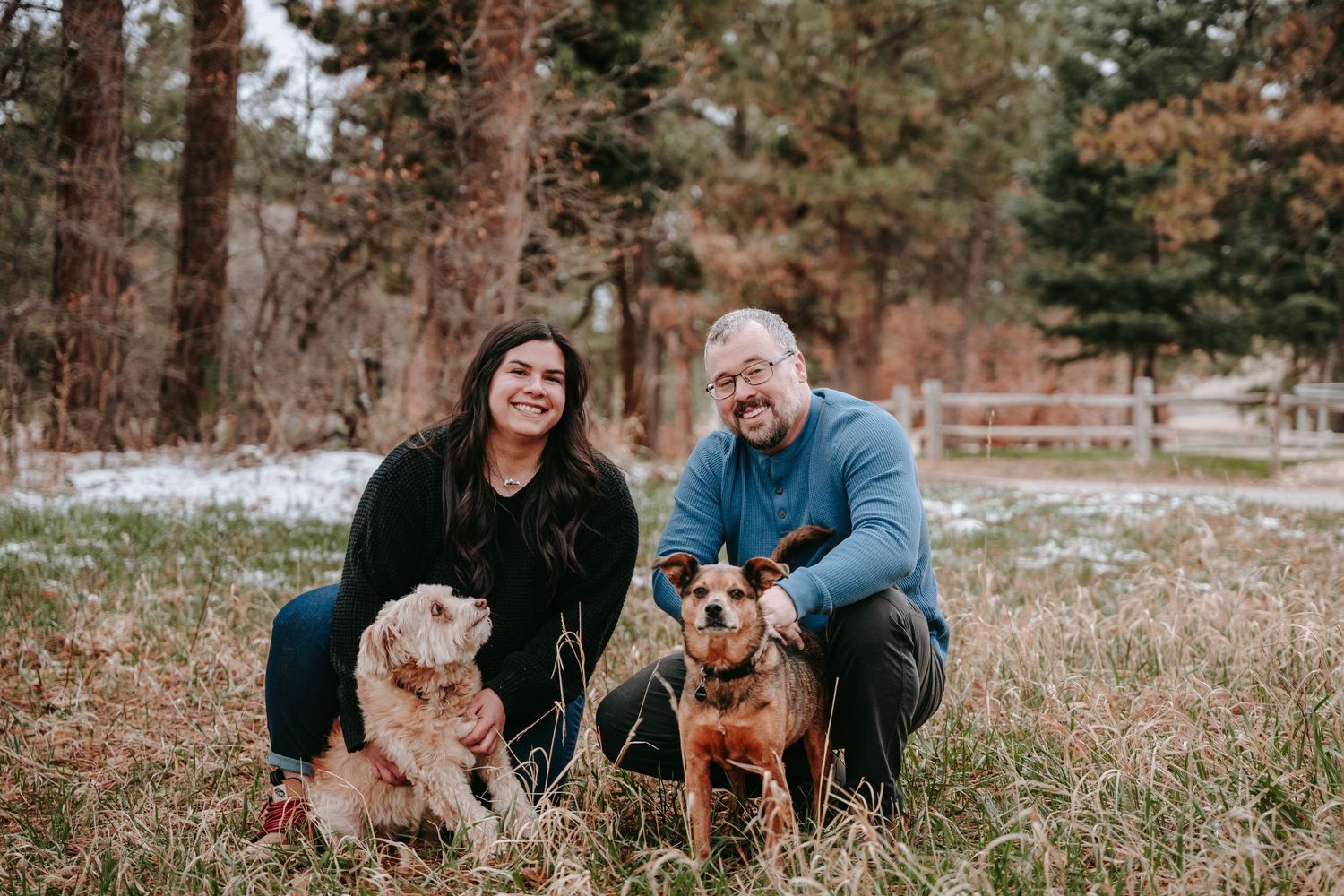 Couple smiling with two dogs in a snowy forest field during winter family pet photo session.