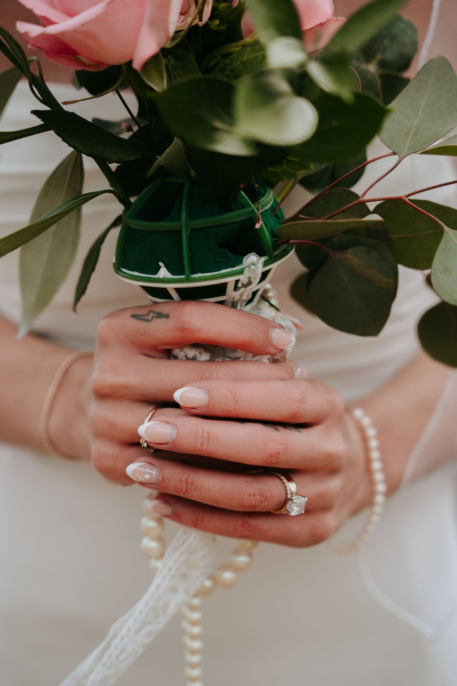 Bride holding pink rose bouquet with diamond ring and pearl bracelet on her manicured hands.