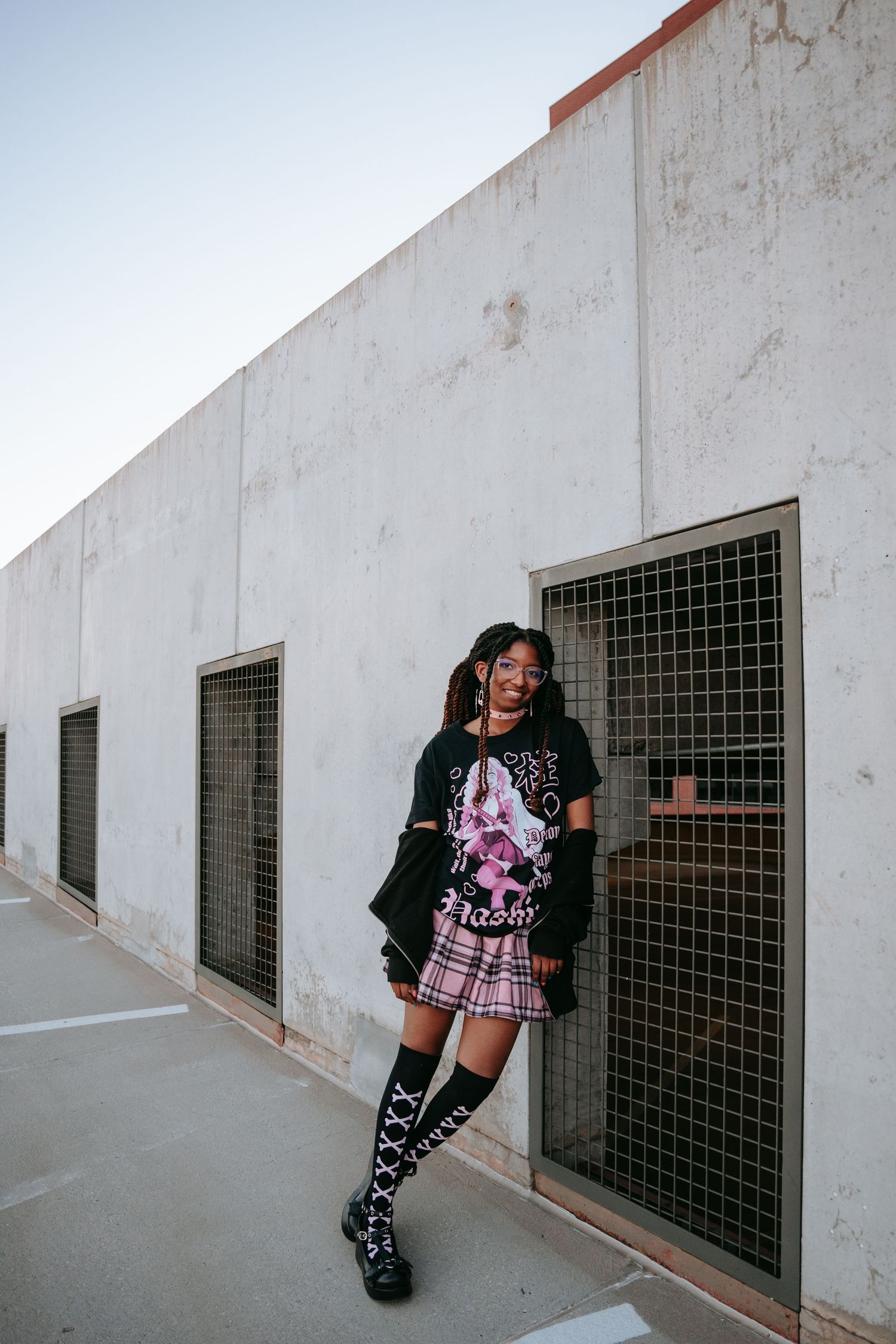 Young woman in edgy outfit with graphic tee, plaid skirt, and knee-high boots leaning against wall.